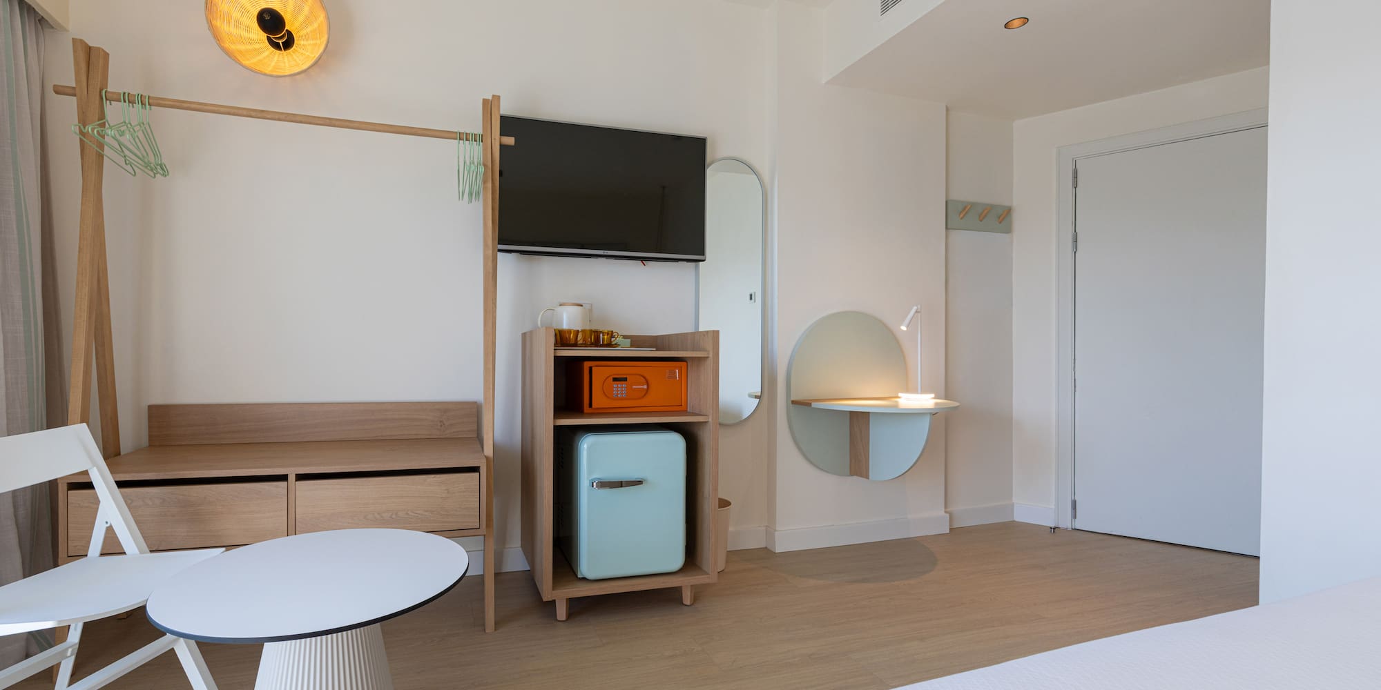 Modern hotel room with light wood furniture, TV, mint minibar, and safe.