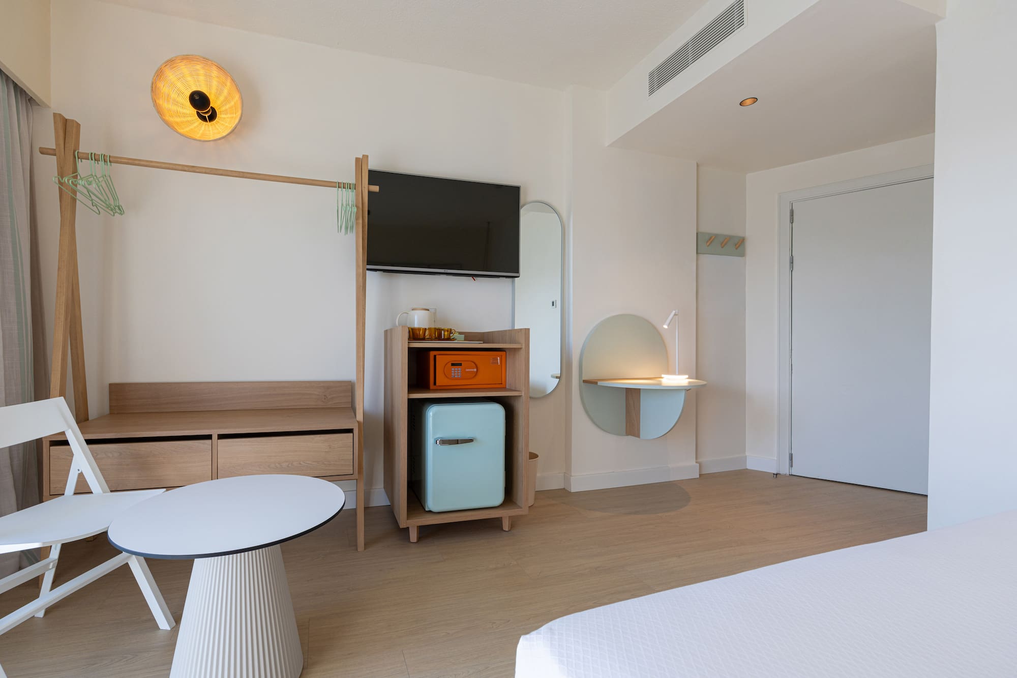 Modern hotel guest room with TV, mini-fridge, wooden furniture, and round desk.