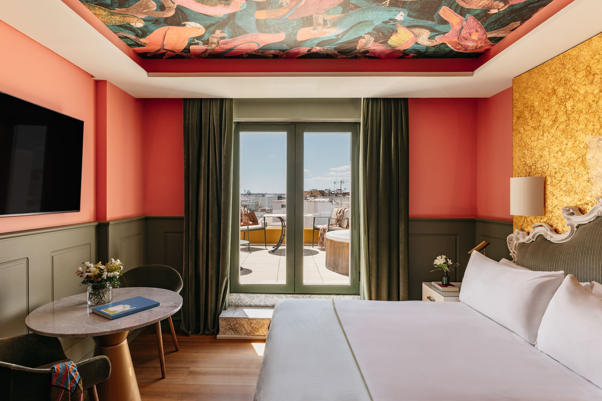 Suite de hotel colorida con mural techo, paredes coral y balcón privado.