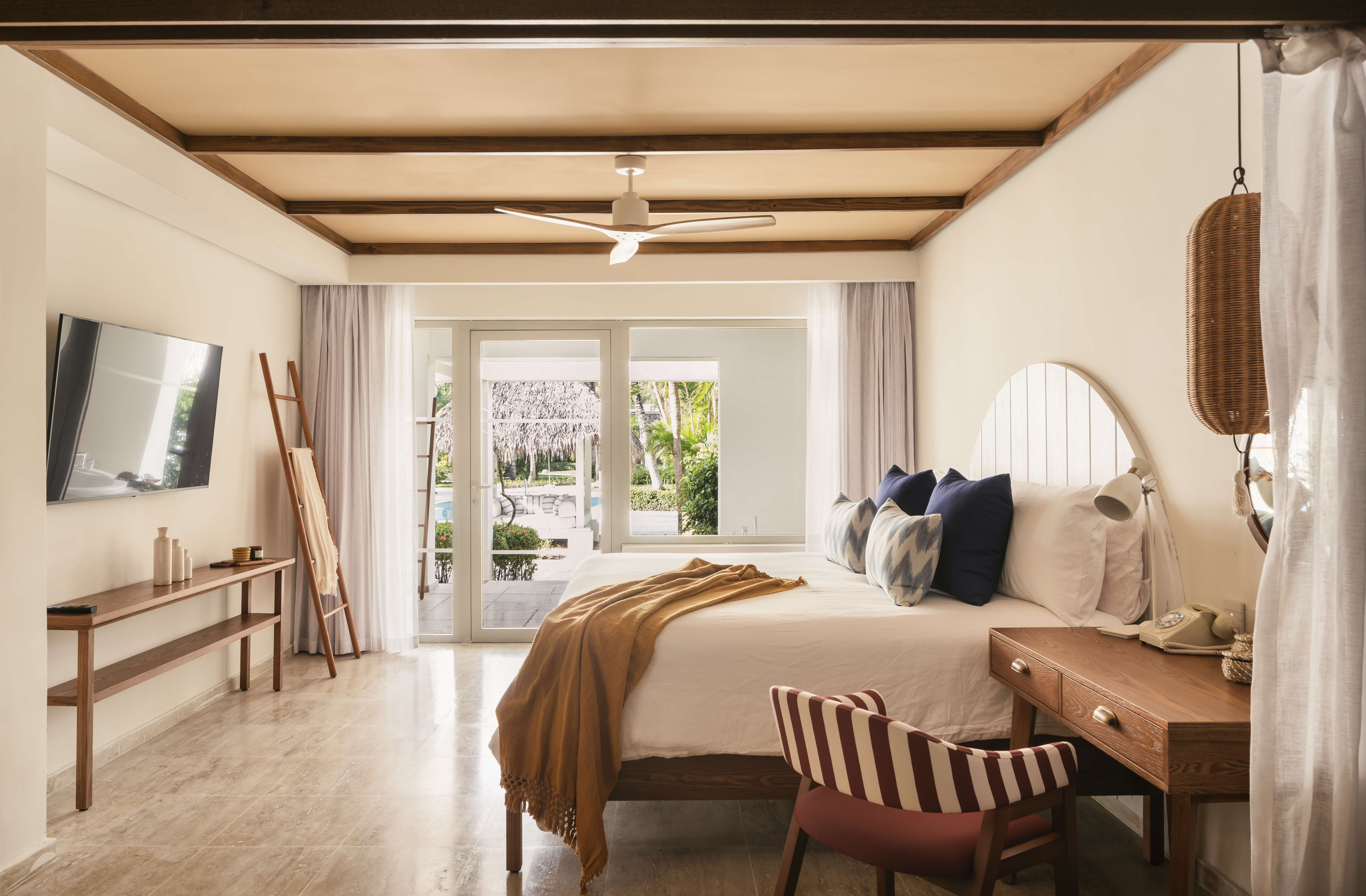 Tropical hotel bedroom with wooden beams, white bed, and pool view.