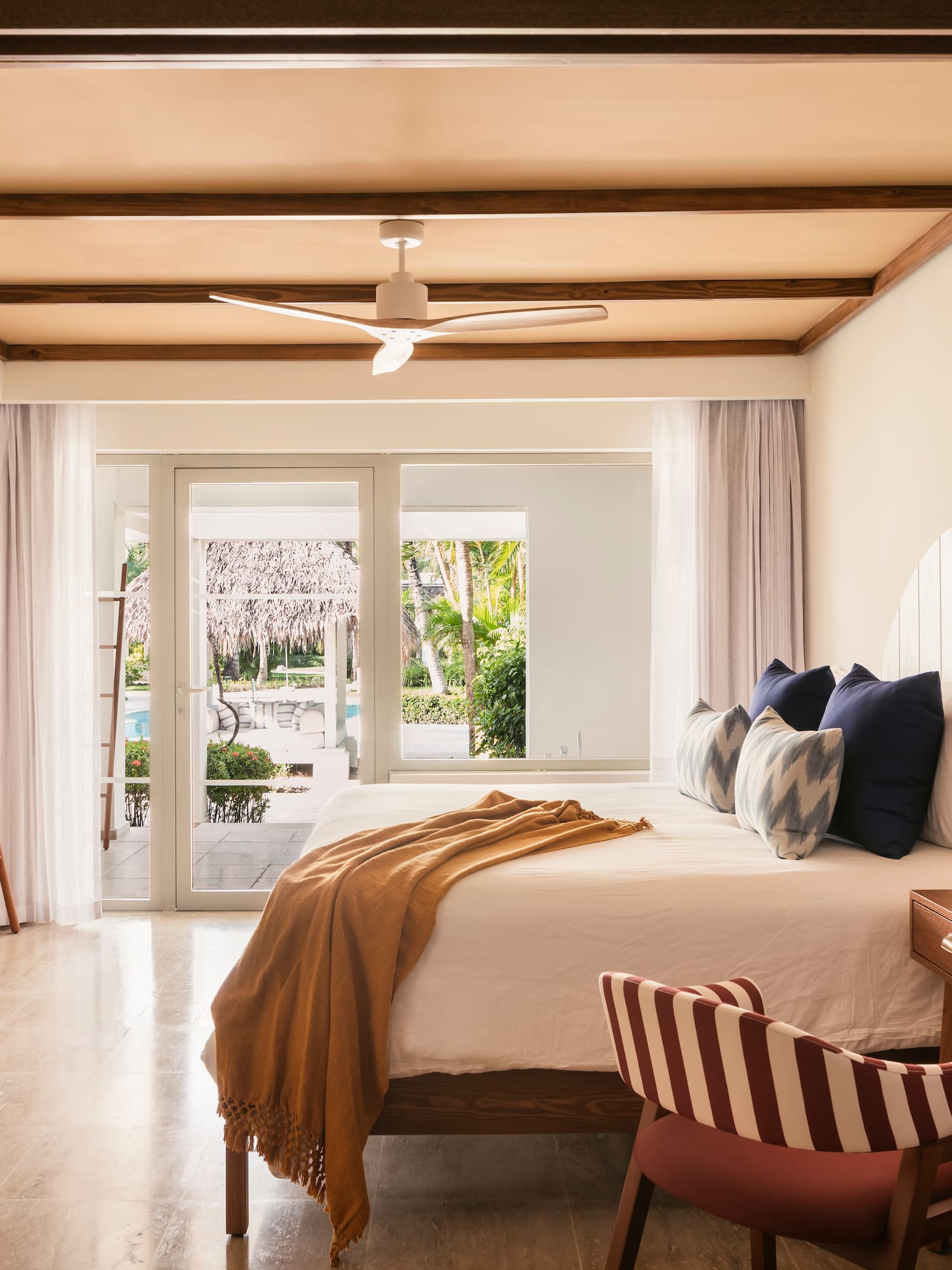 Modern tropical bedroom with bed, wooden desk, TV, and outdoor patio view.