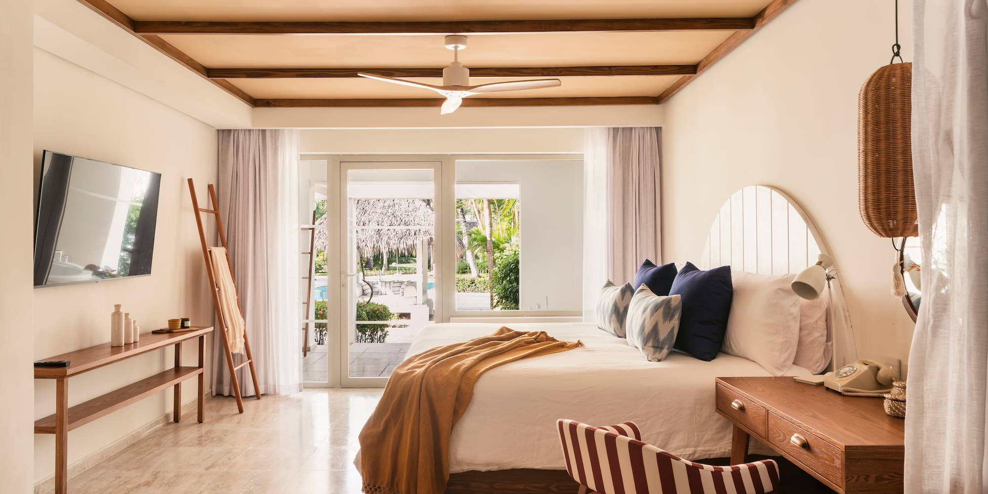 Tropical hotel bedroom with wooden beams, white bed, and pool view.