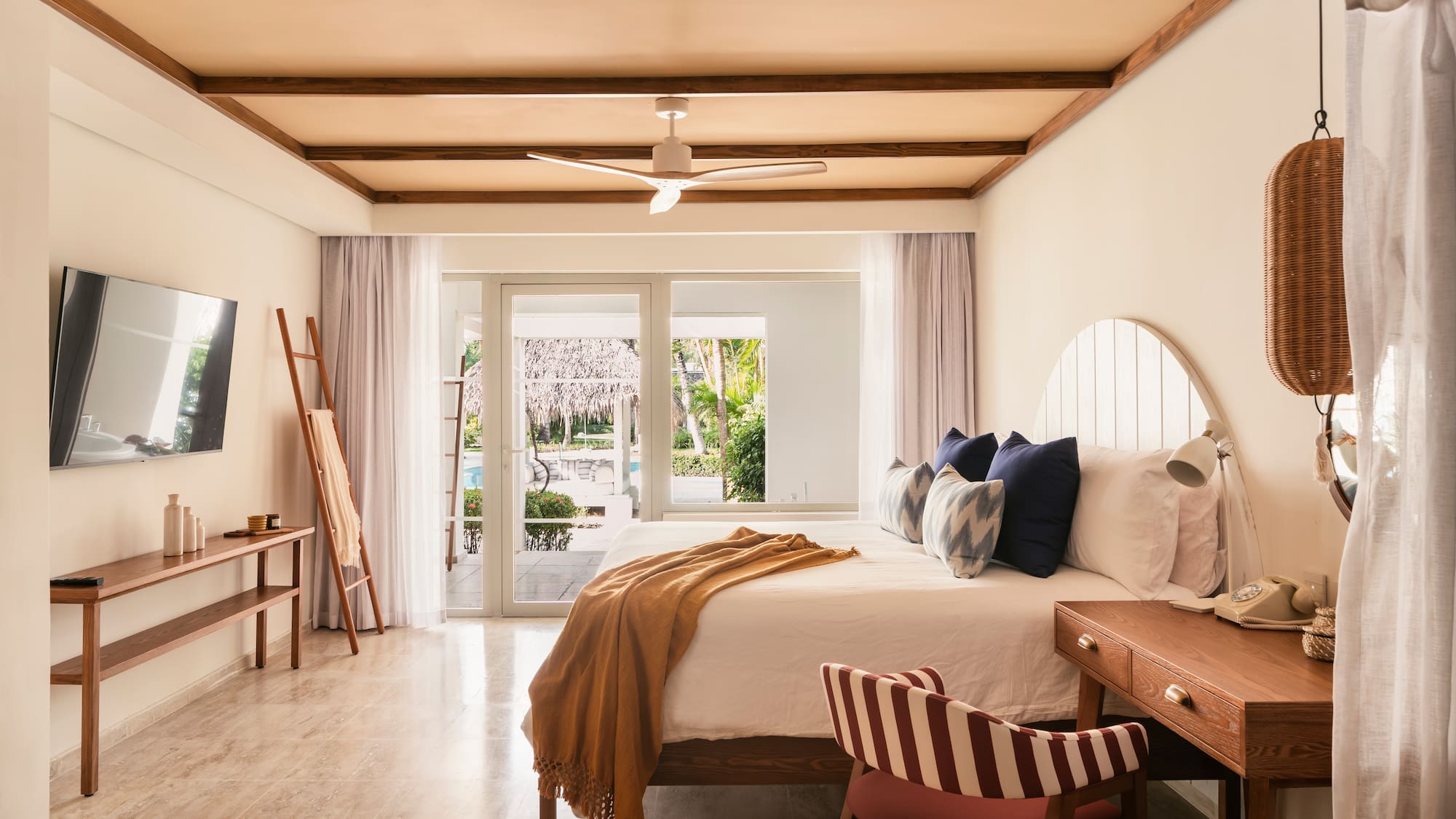 Tropical hotel bedroom with wooden beams, white bed, and pool view.