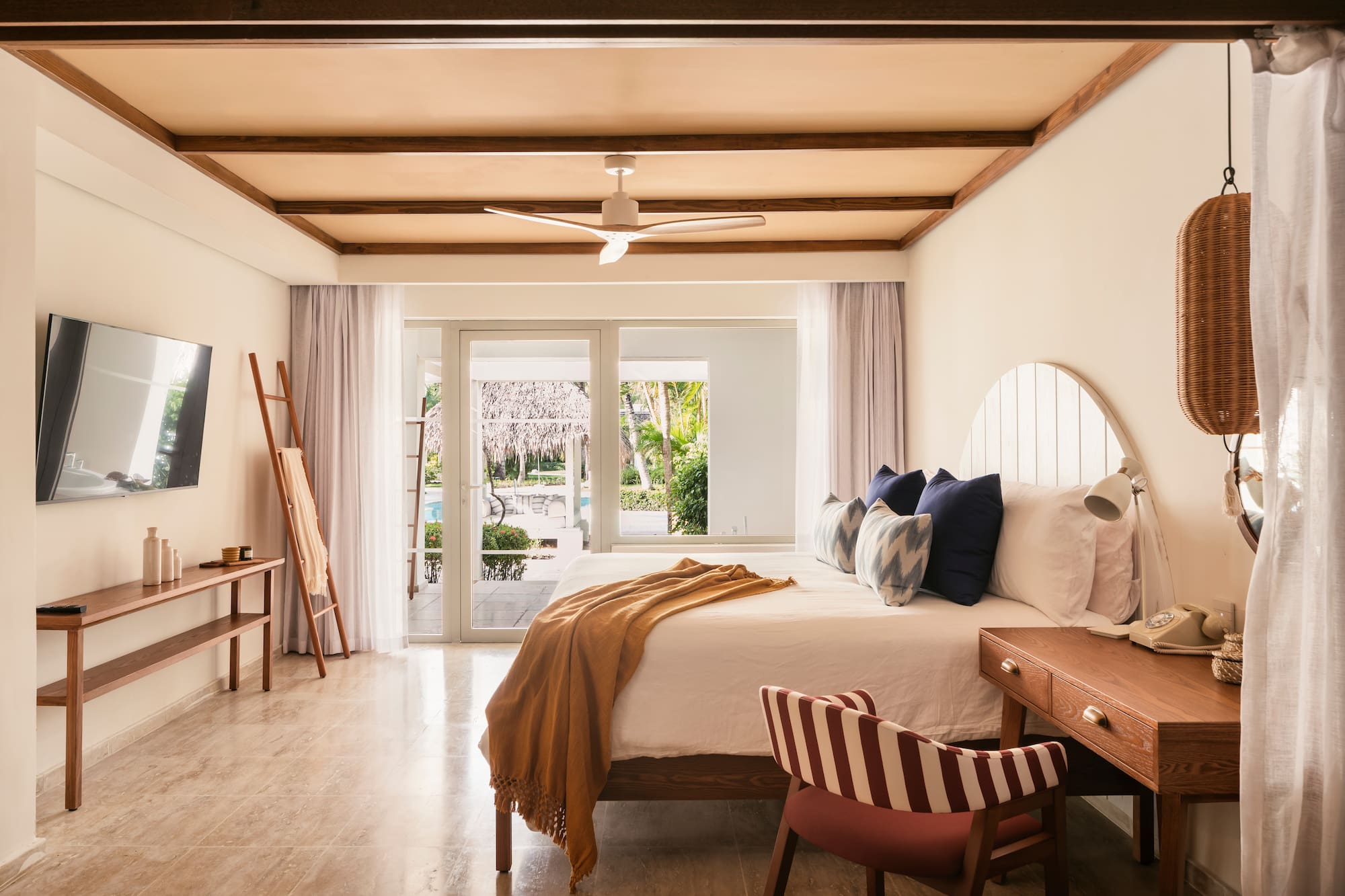 Tropical hotel bedroom with wooden beams, white bed, and pool view.