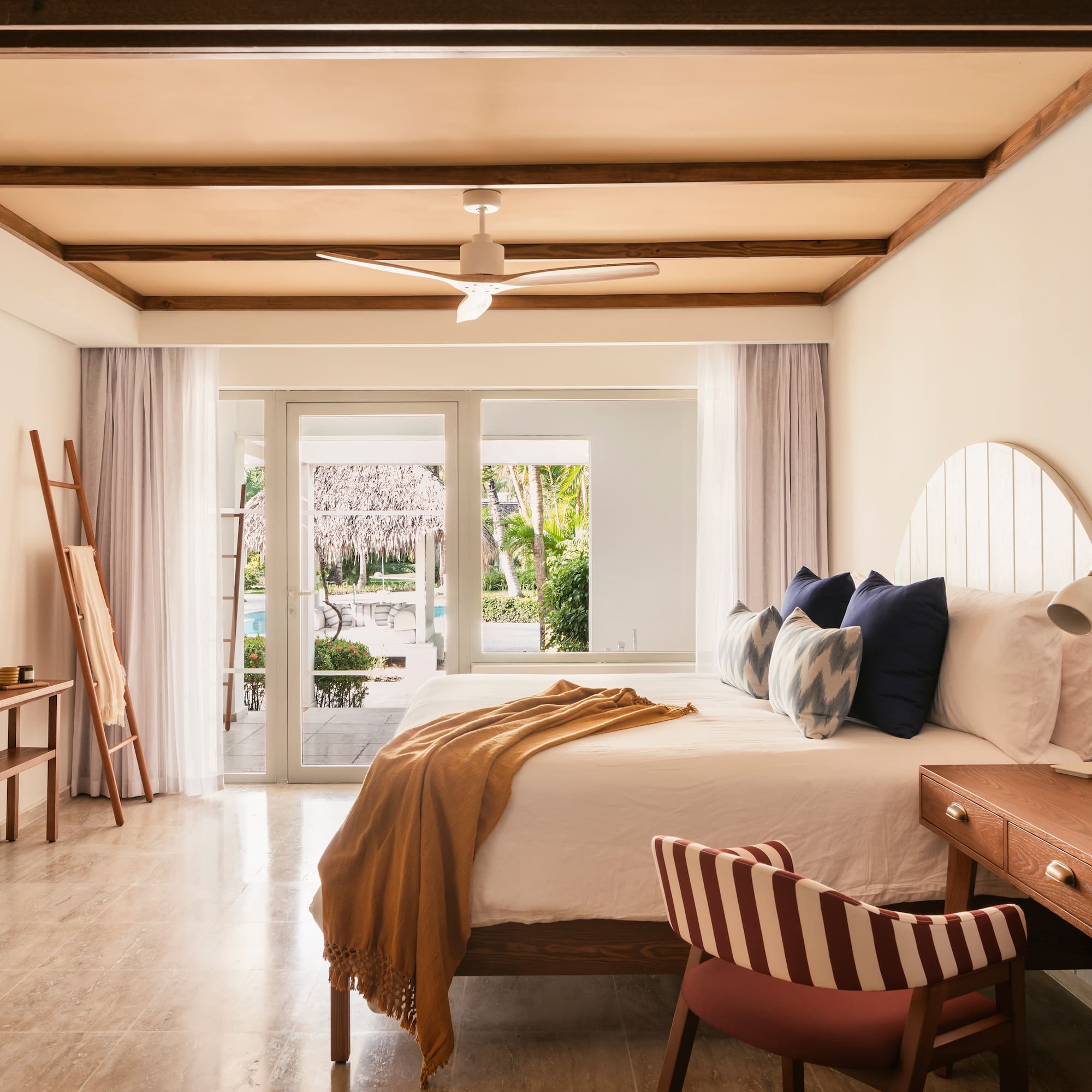 Tropical hotel bedroom with wooden beams, white bed, and pool view.