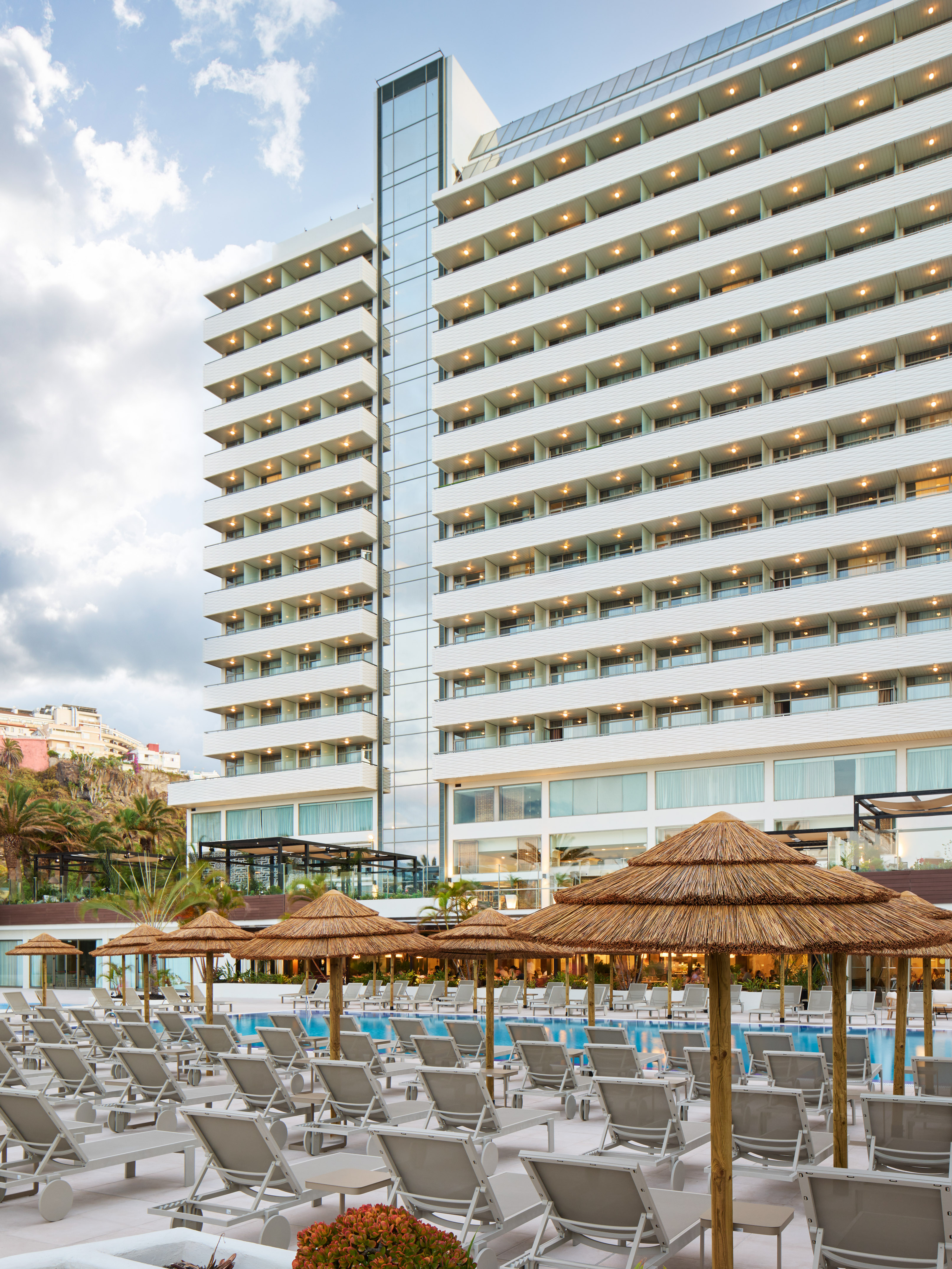Modern hotel with pool, sun loungers, straw parasols, and bright balconies.