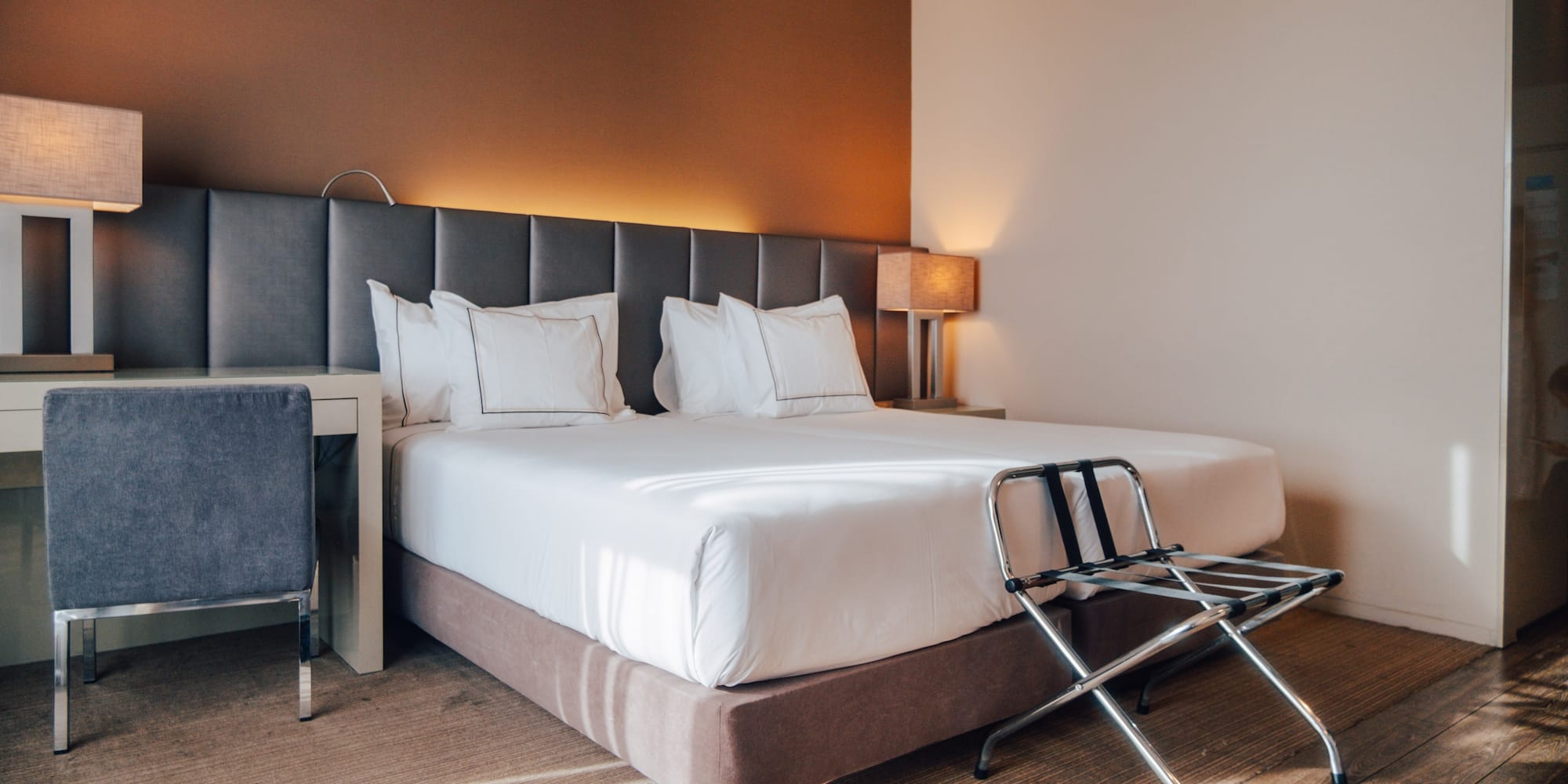 Modern hotel bedroom with large bed, grey headboard, desk, and luggage rack.