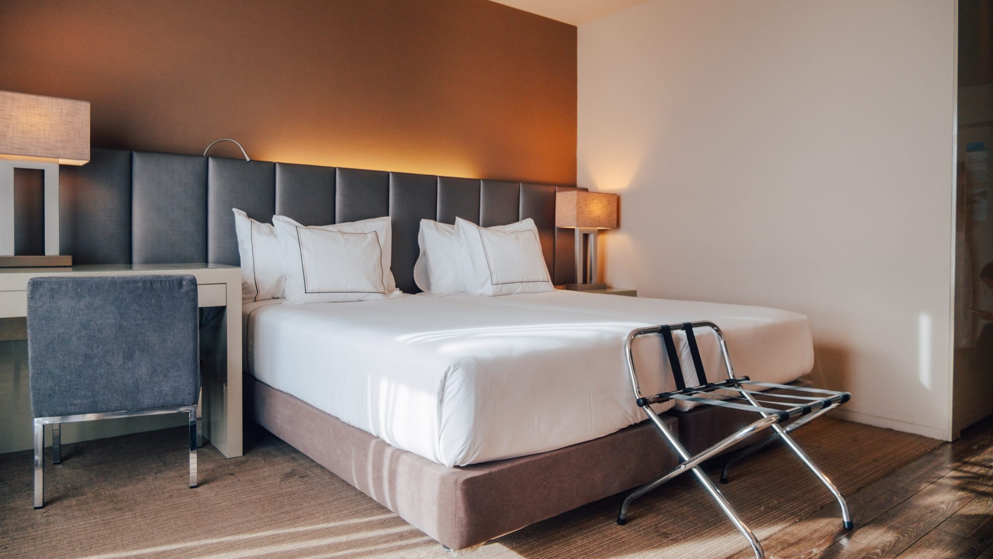 Modern hotel bedroom with large bed, grey headboard, desk, and luggage rack.