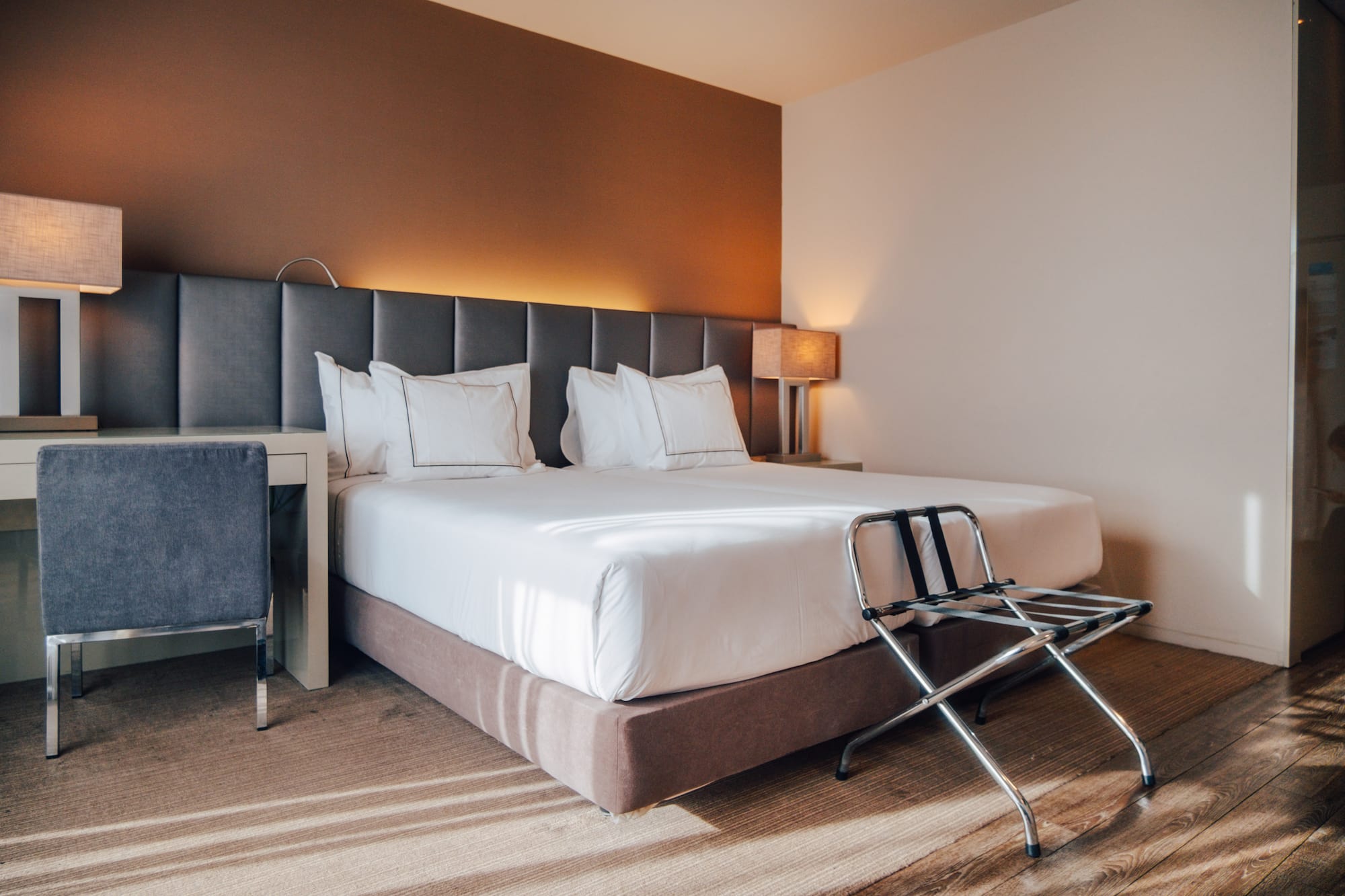 Modern hotel bedroom with large bed, grey headboard, desk, and luggage rack.