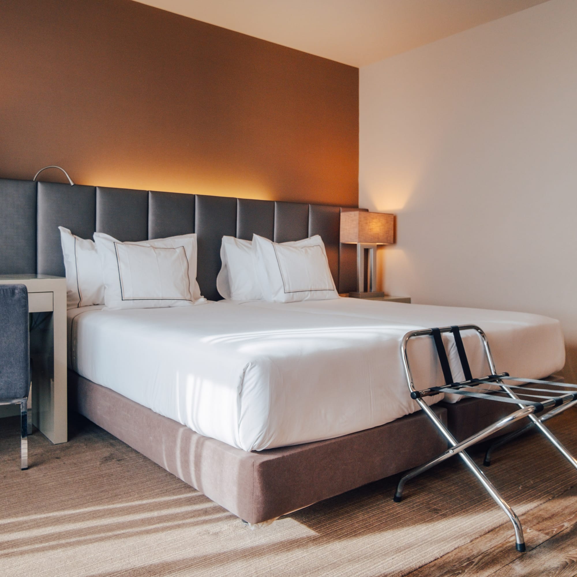 Modern hotel bedroom with large bed, grey headboard, desk, and luggage rack.