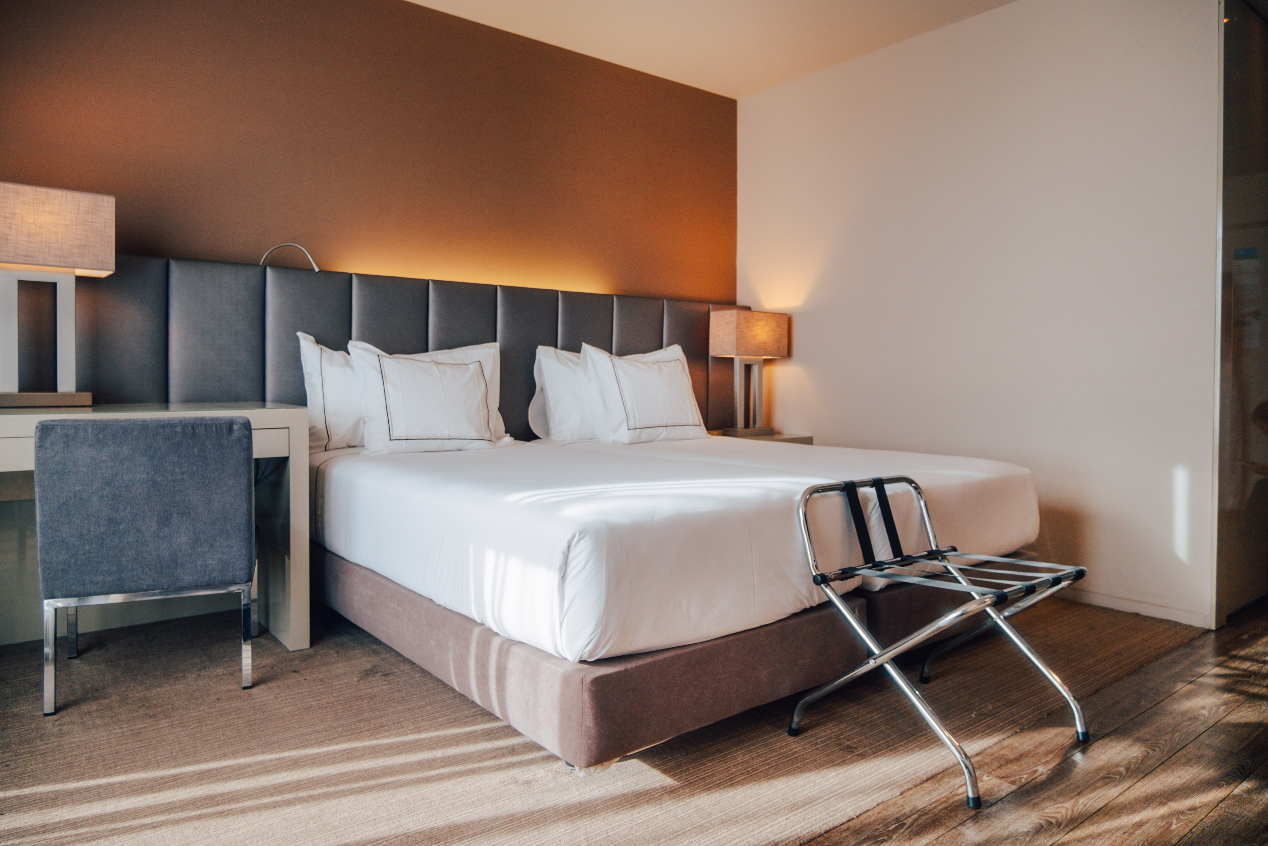 Modern hotel bedroom with large bed, grey headboard, desk, and luggage rack.