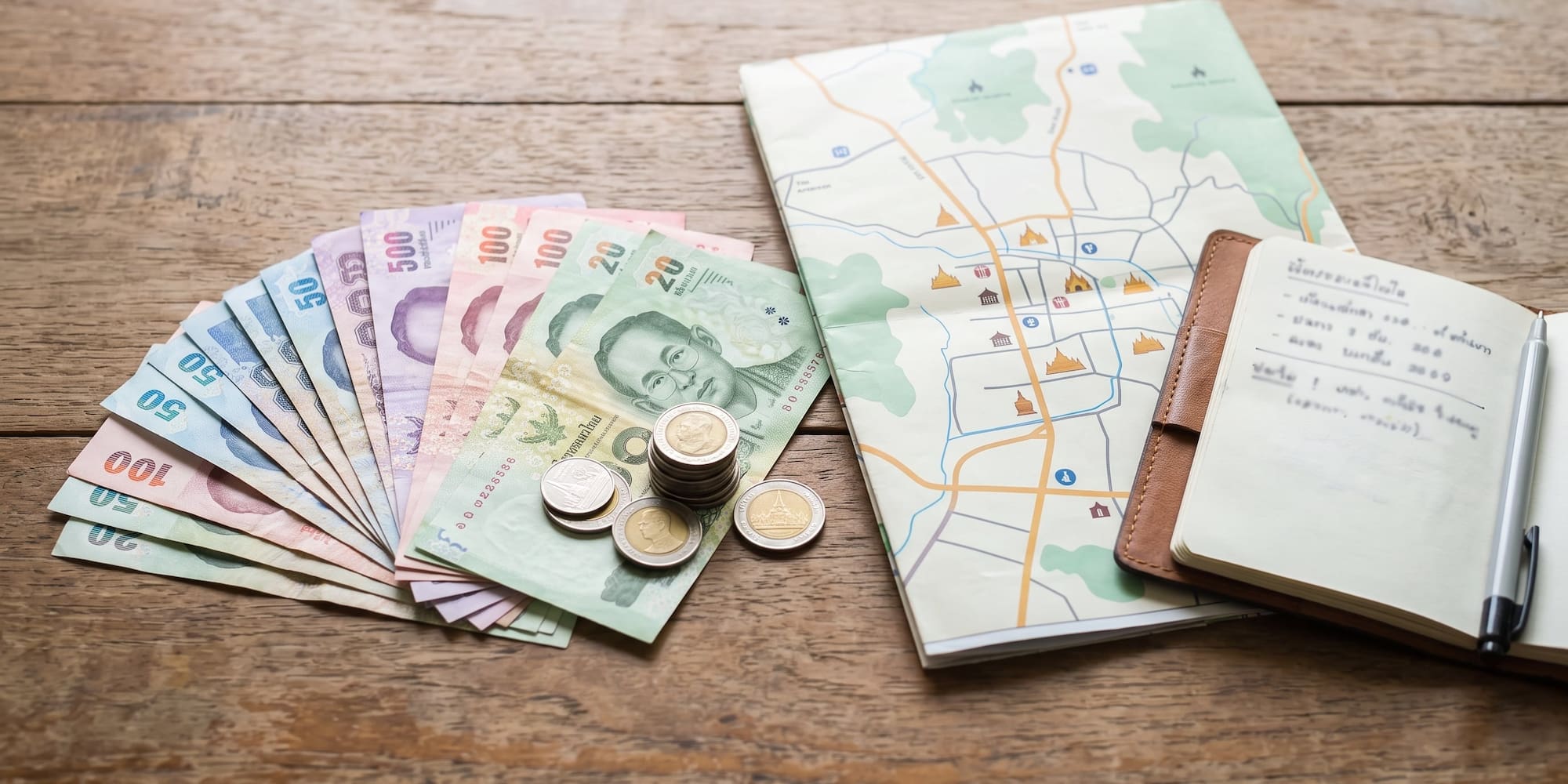 Thai Baht cash, coins, travel map, and notebook on wood. Trip planning.