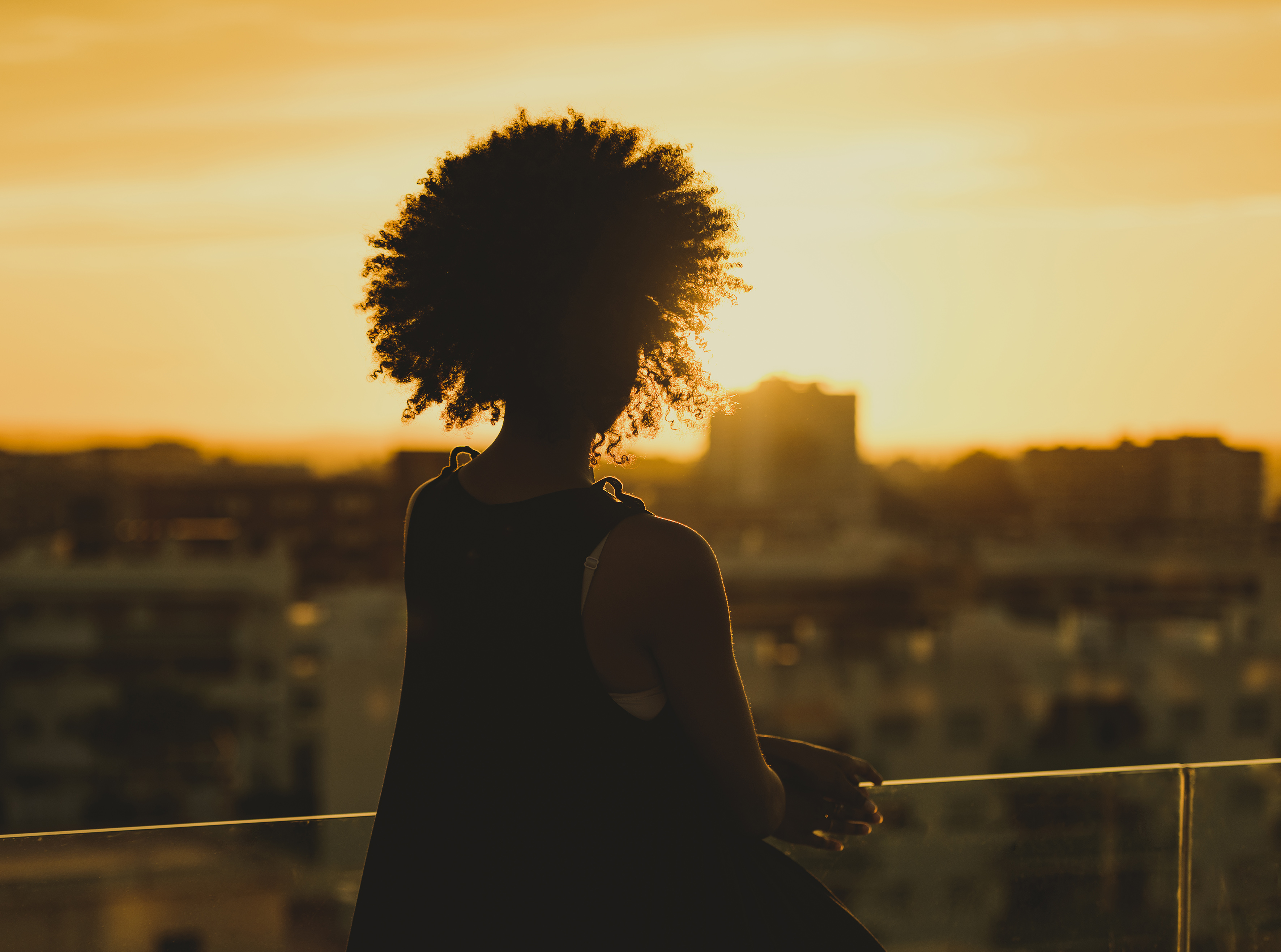 Silhouetted person with curly hair watching a golden sunset over a city skyline.