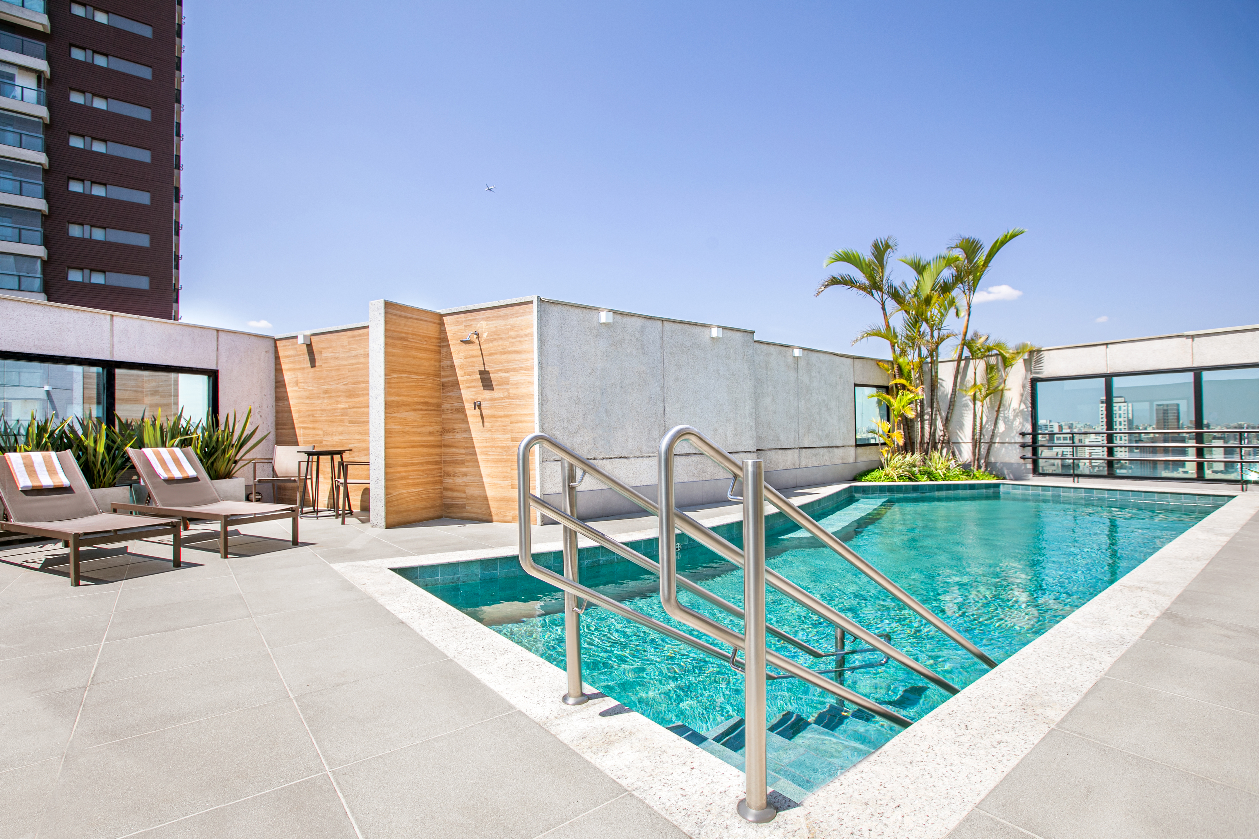 Modern rooftop pool deck with blue water, lounge chairs, palm trees, and city skyline.