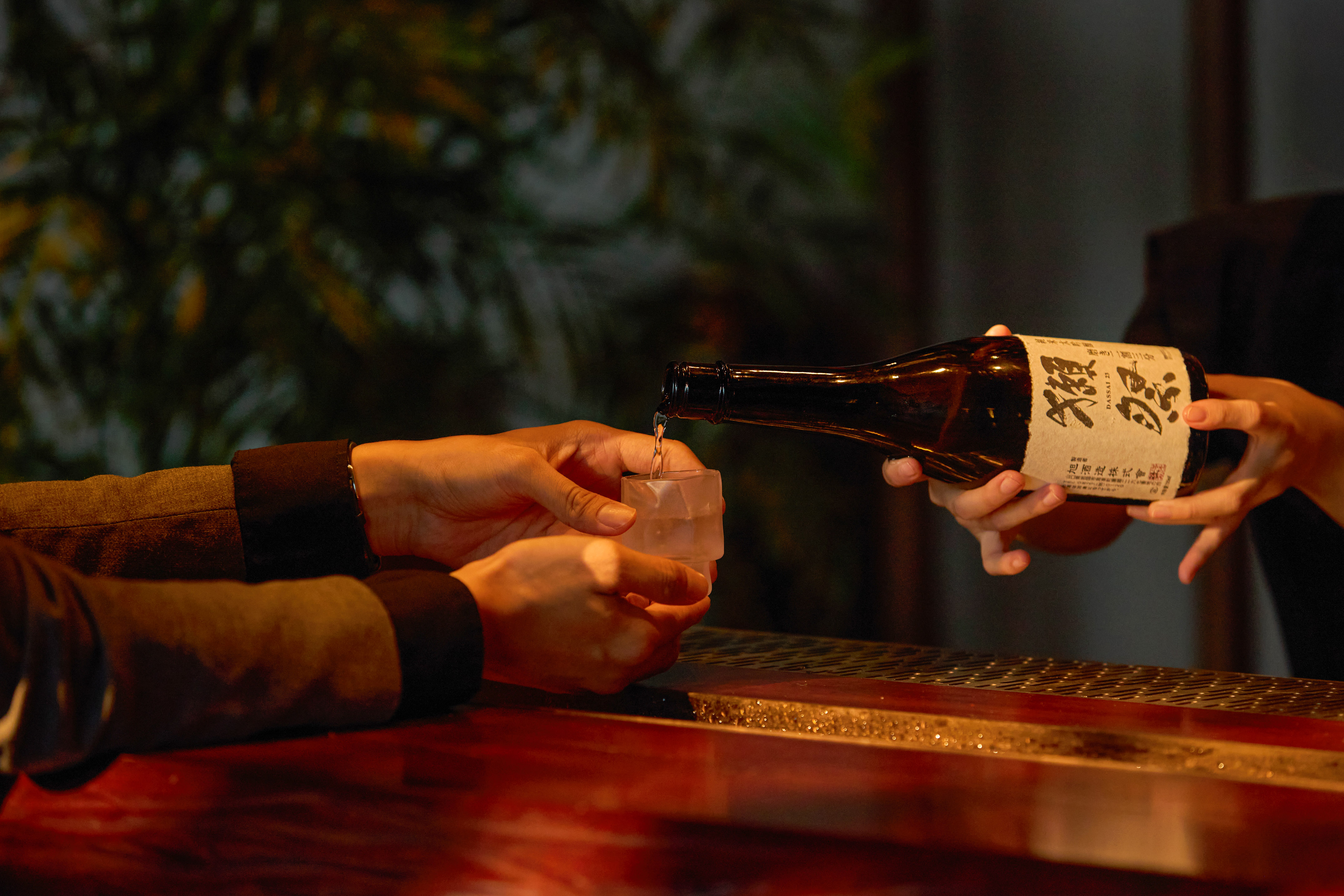 Hands pouring Dassai 23 sake from a bottle into a small glass on a bar counter.