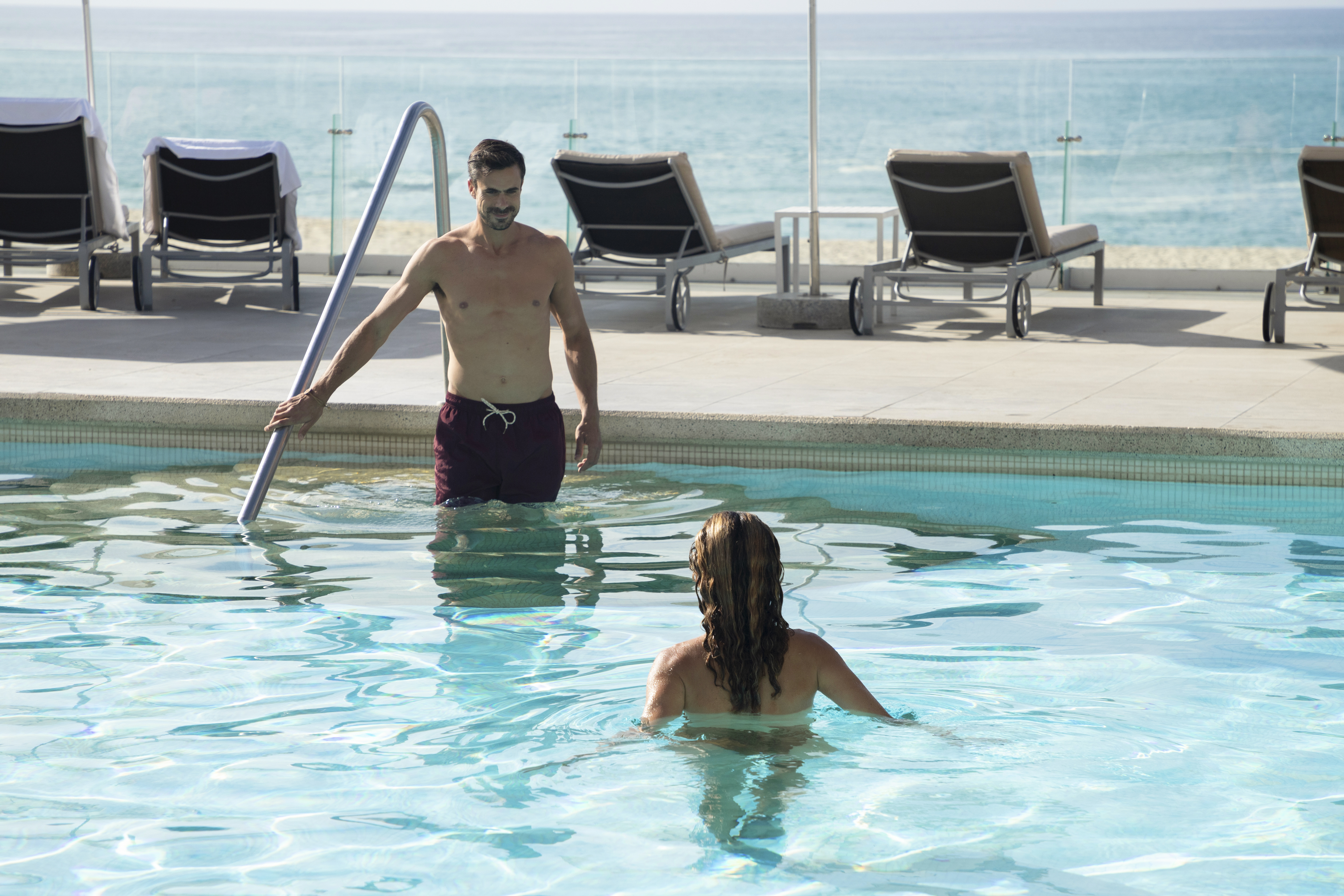 Man exiting resort pool, woman swimming, ocean and lounge chairs in background.