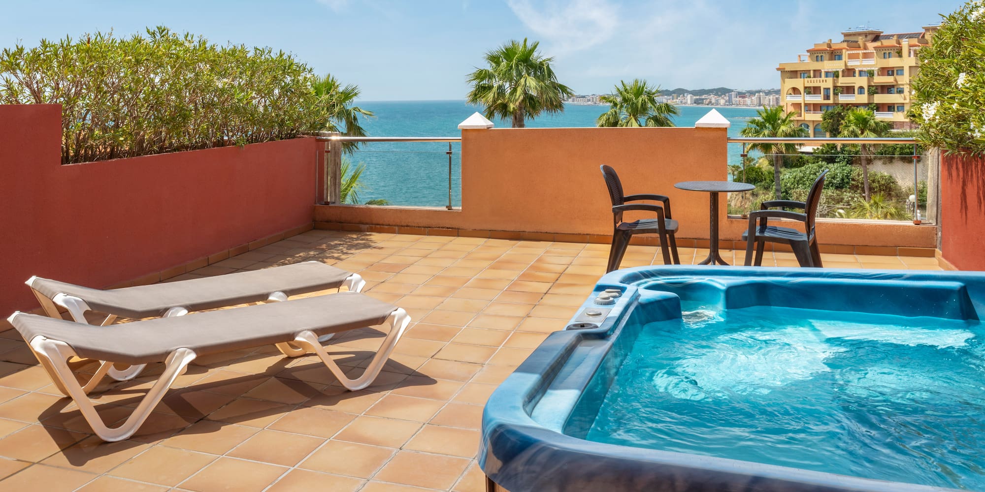 Coastal terrace with hot tub, sun loungers, palm trees, and sea view.