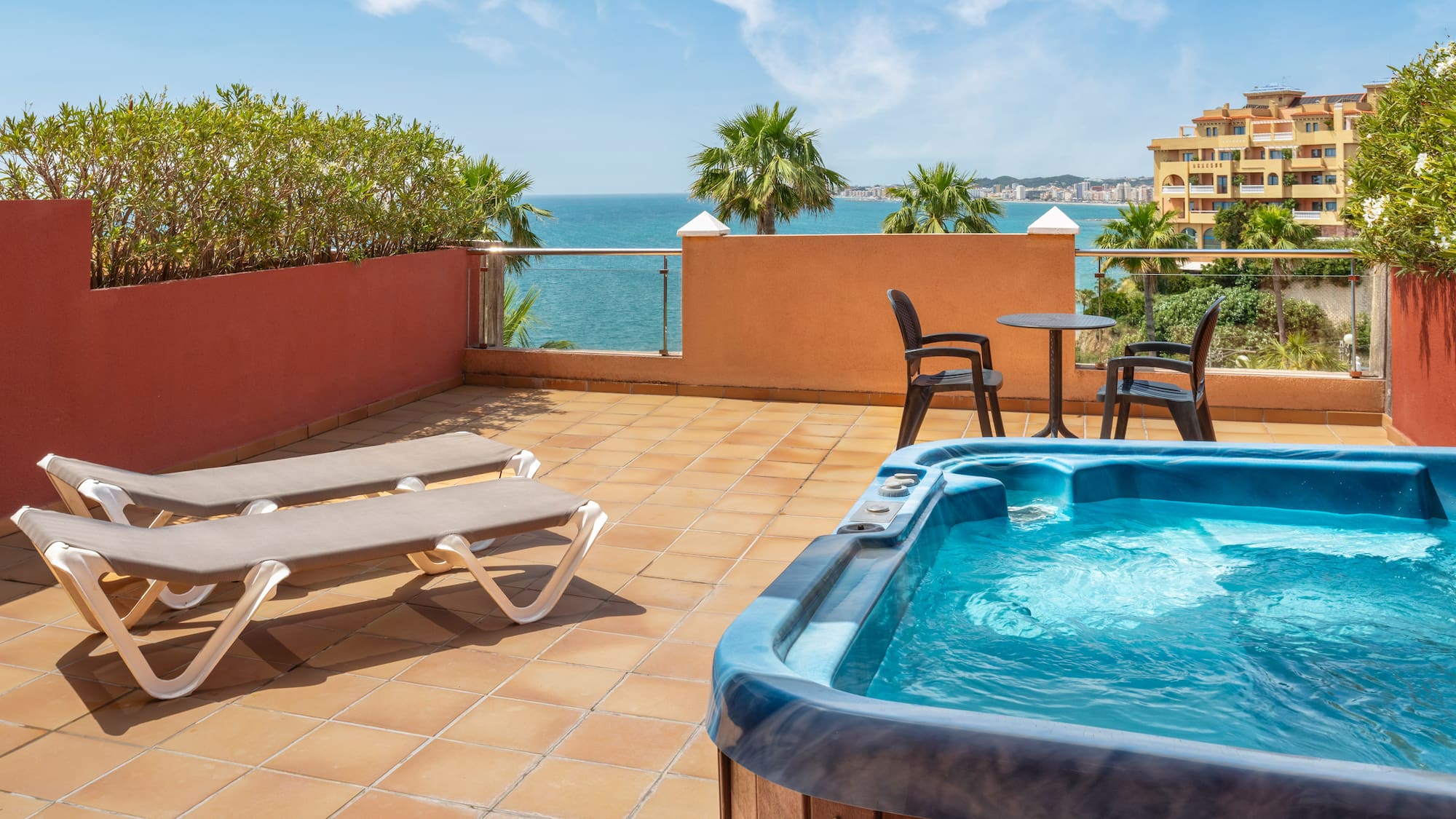 Coastal terrace with hot tub, sun loungers, palm trees, and sea view.