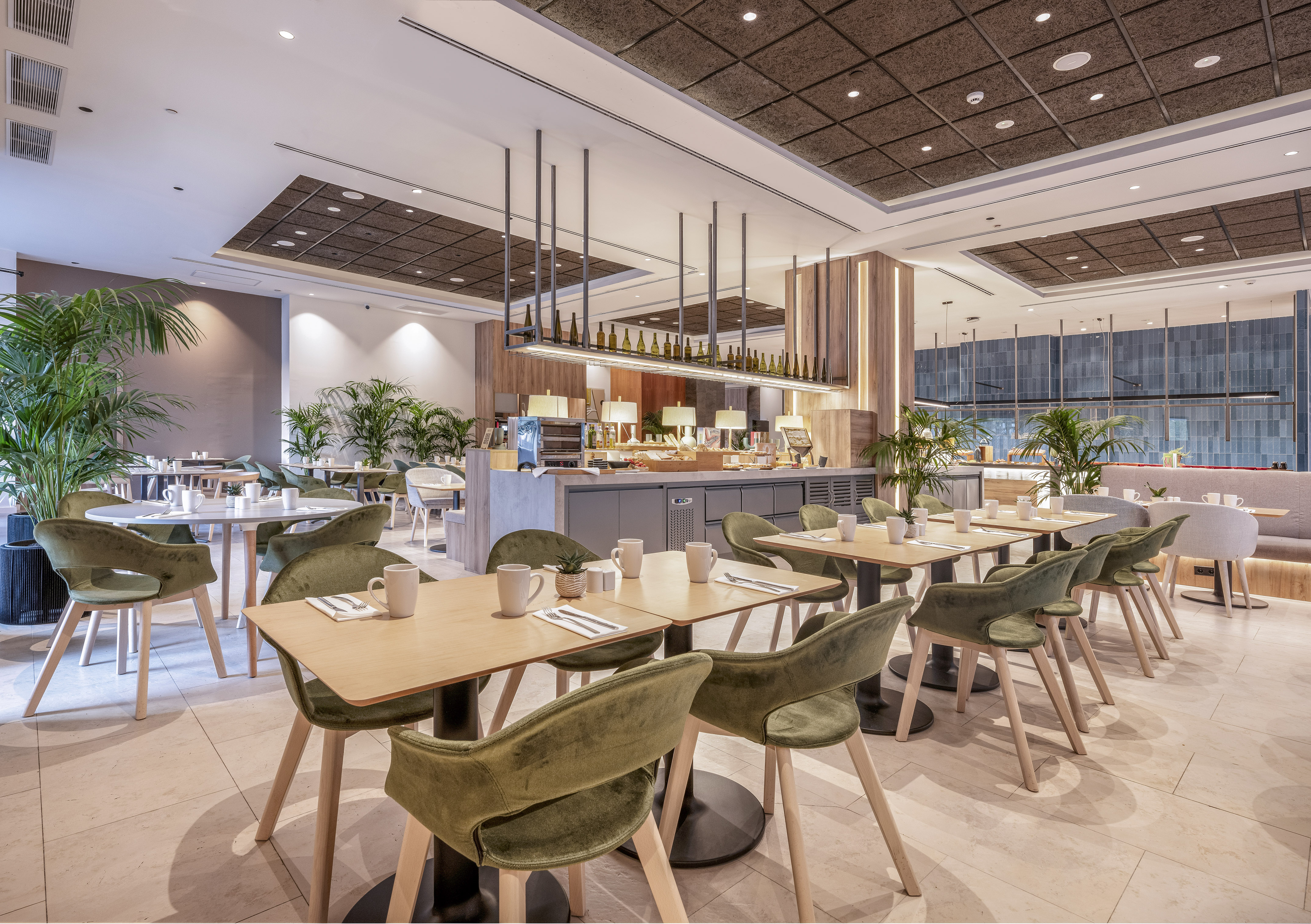 Modern hotel restaurant with green velvet chairs, wooden tables, and buffet.