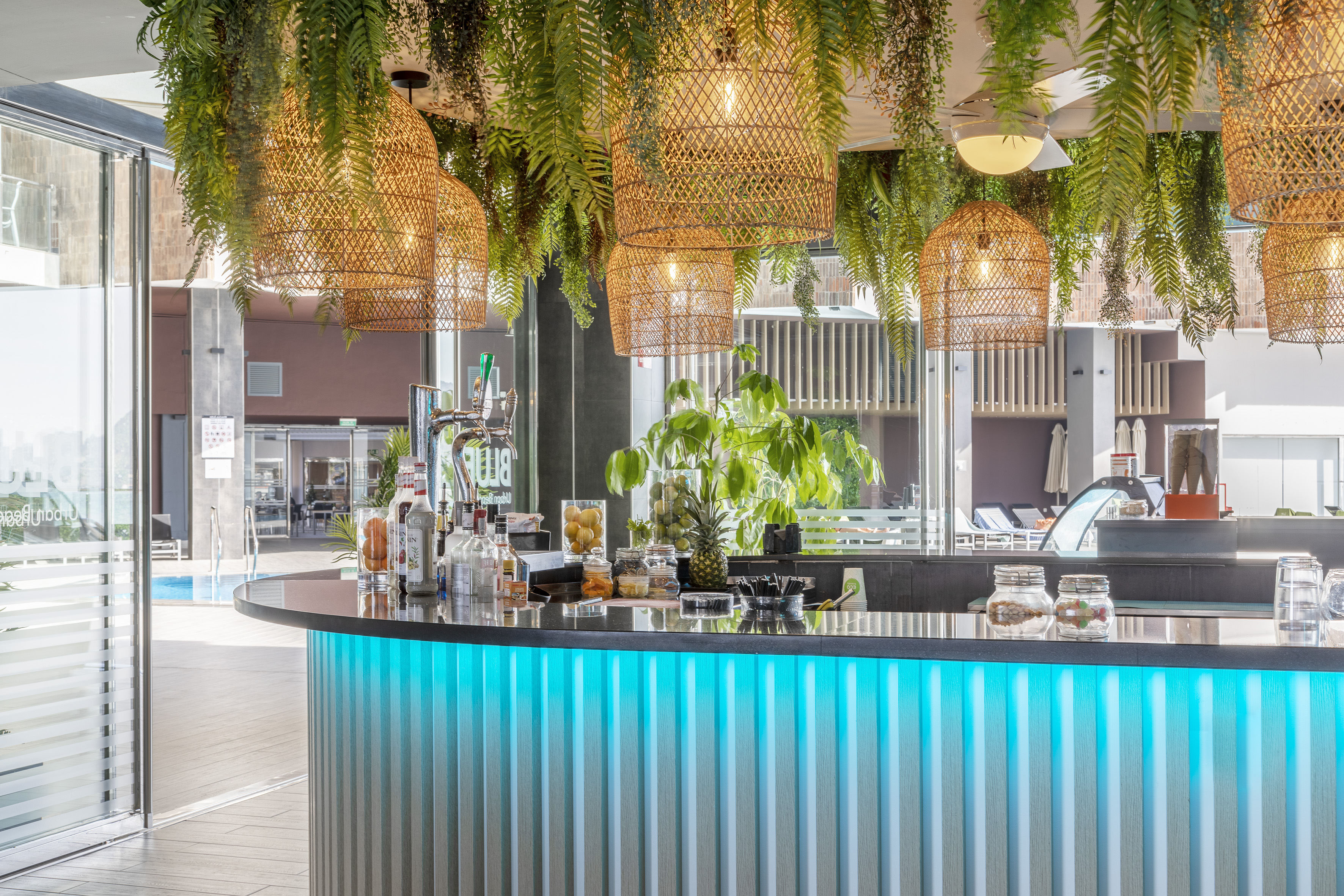 Blue LED resort bar with tropical hanging plants, wicker lights, and pool view.