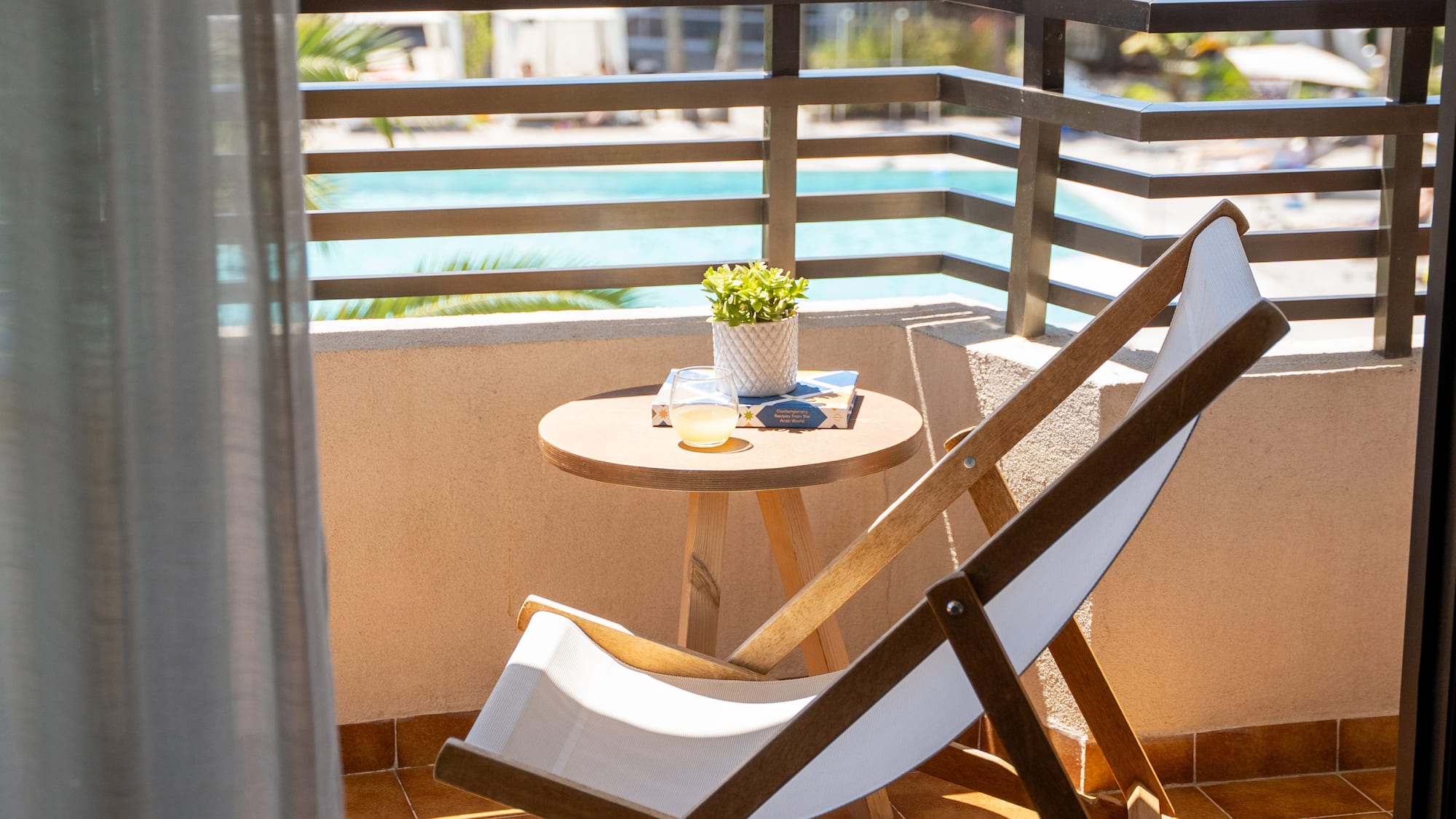 Sunny hotel balcony with deck chair, table, plant, drink, and pool view.