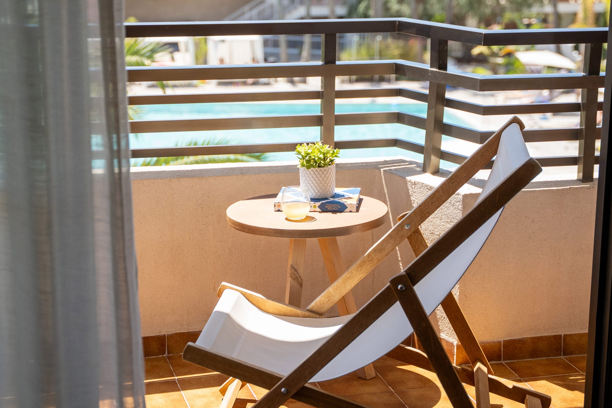 Sunny hotel balcony with deck chair, table, plant, drink, and pool view.