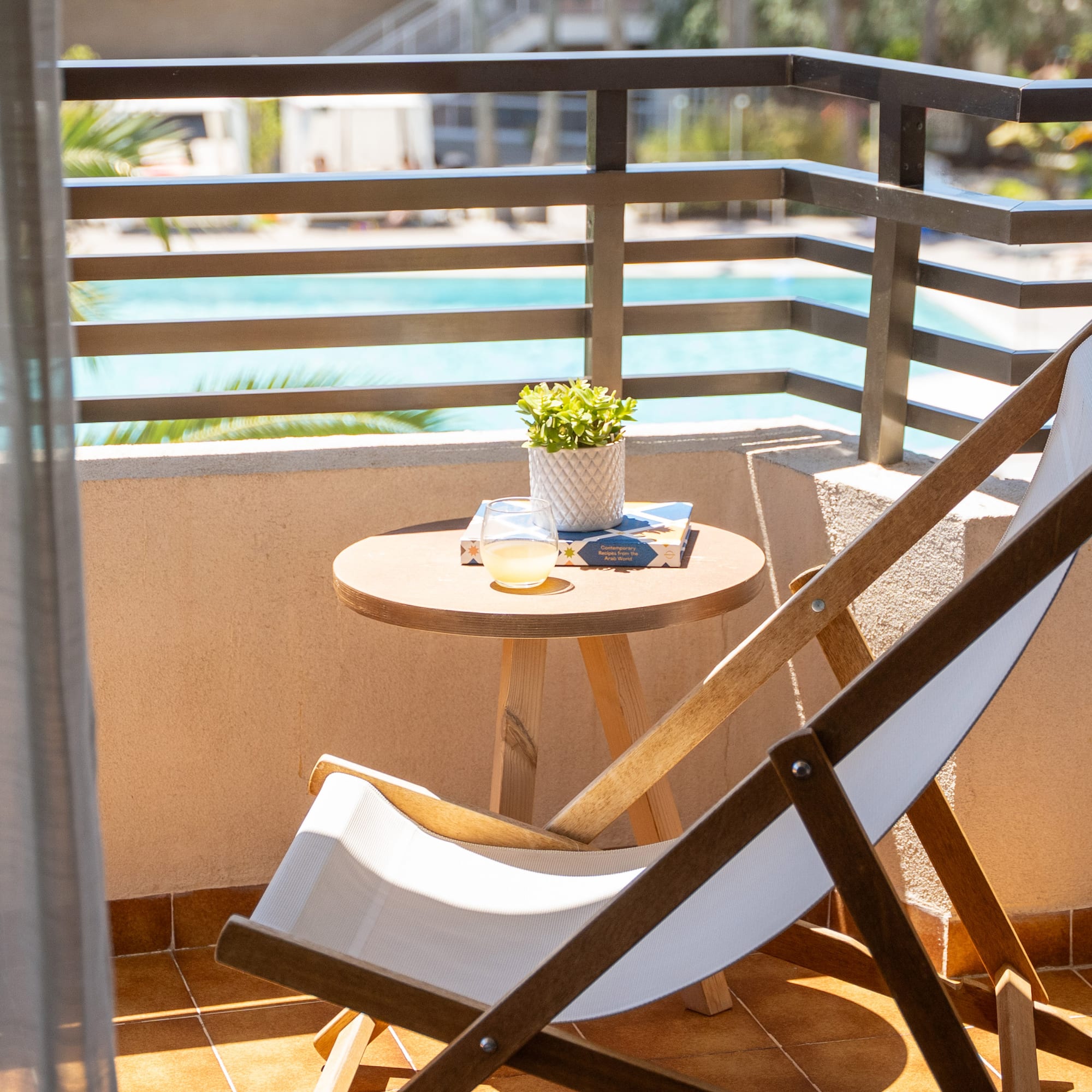 Sunny hotel balcony with deck chair, table, plant, drink, and pool view.