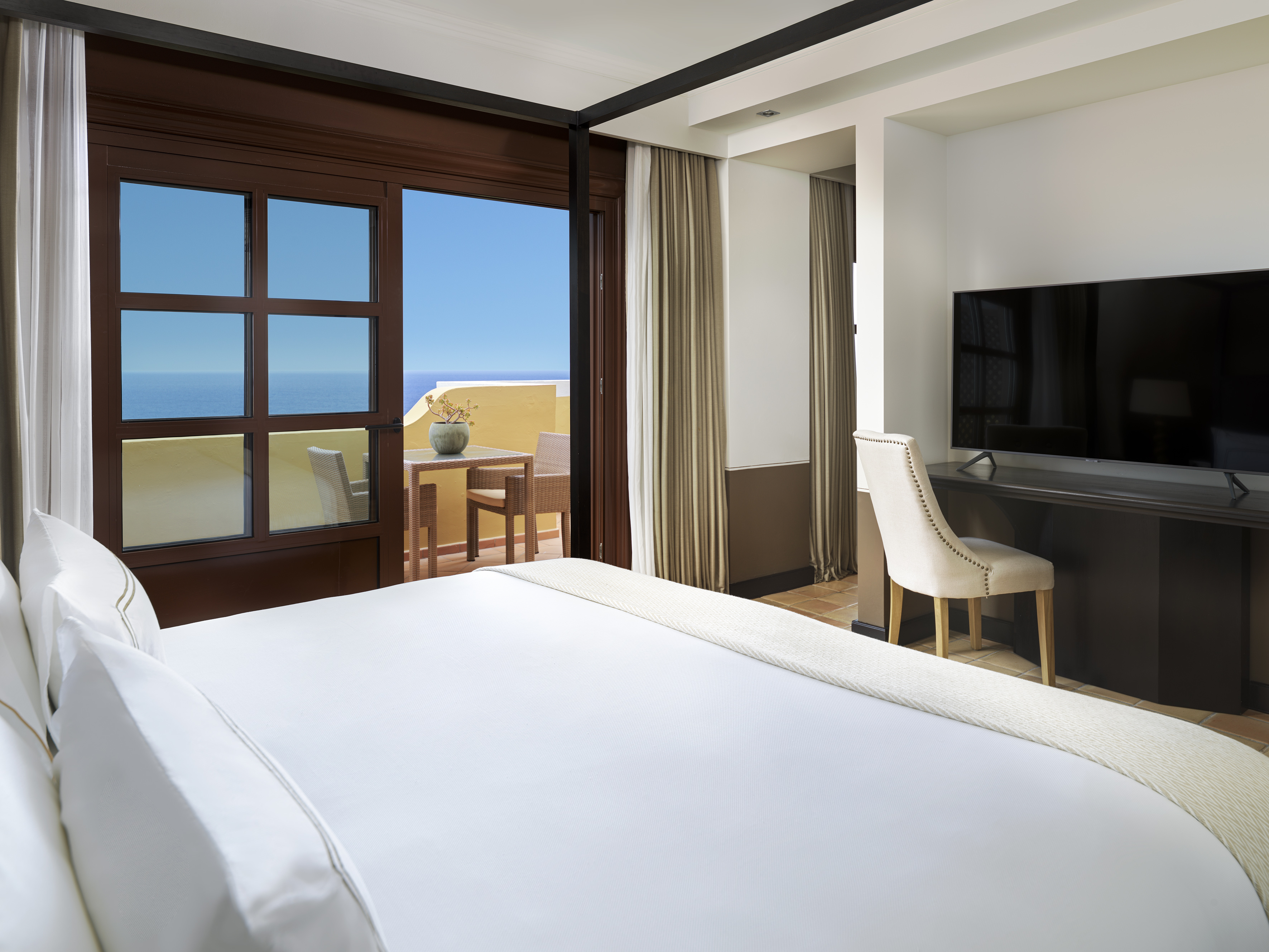 Hotel bedroom with open balcony, ocean view, white bed, TV, and desk.