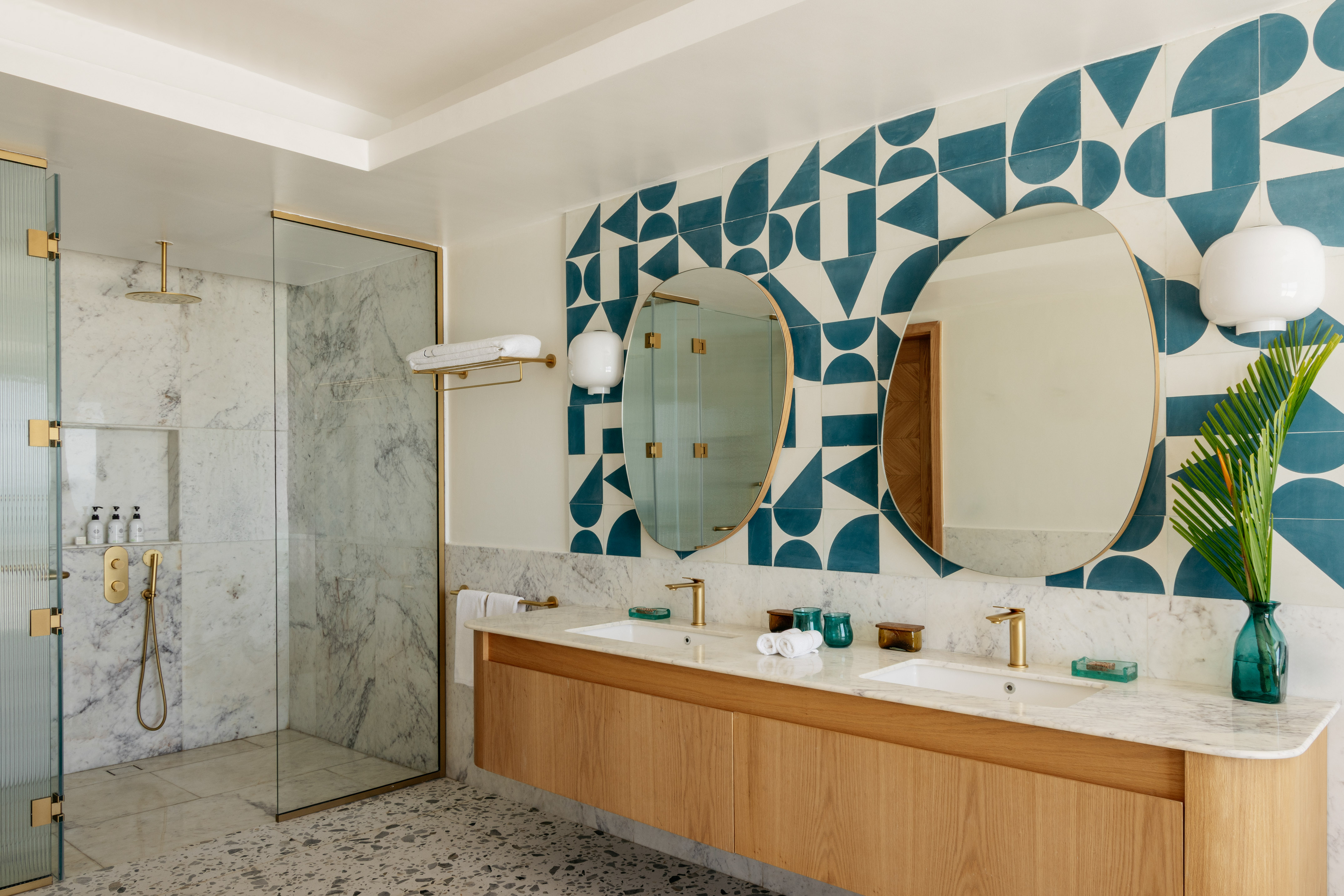 Modern bathroom with blue geometric tiles, wood double vanity, marble shower, and gold fixtures.