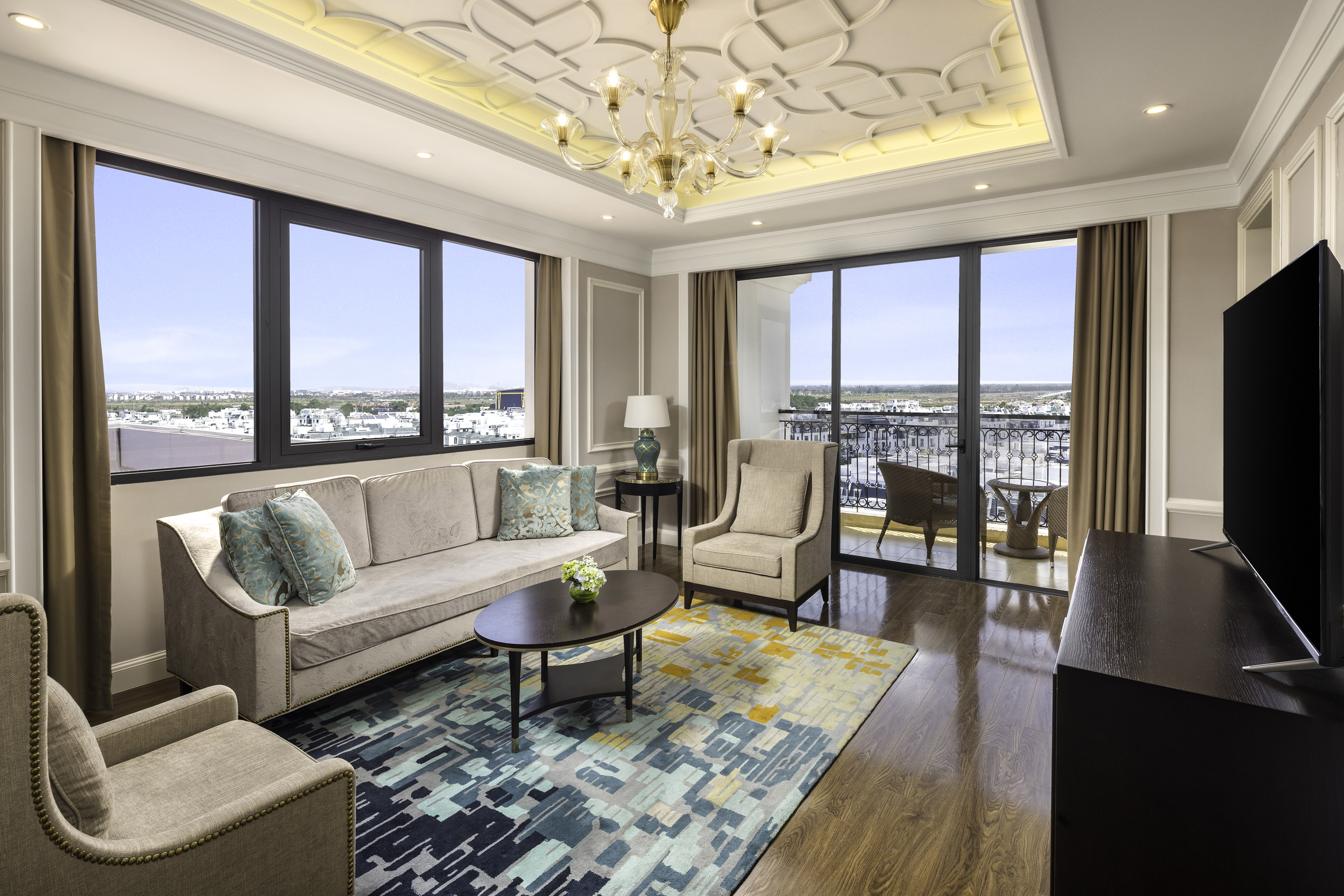 Luxury hotel suite with city view, balcony, modern furnishings, and chandelier.