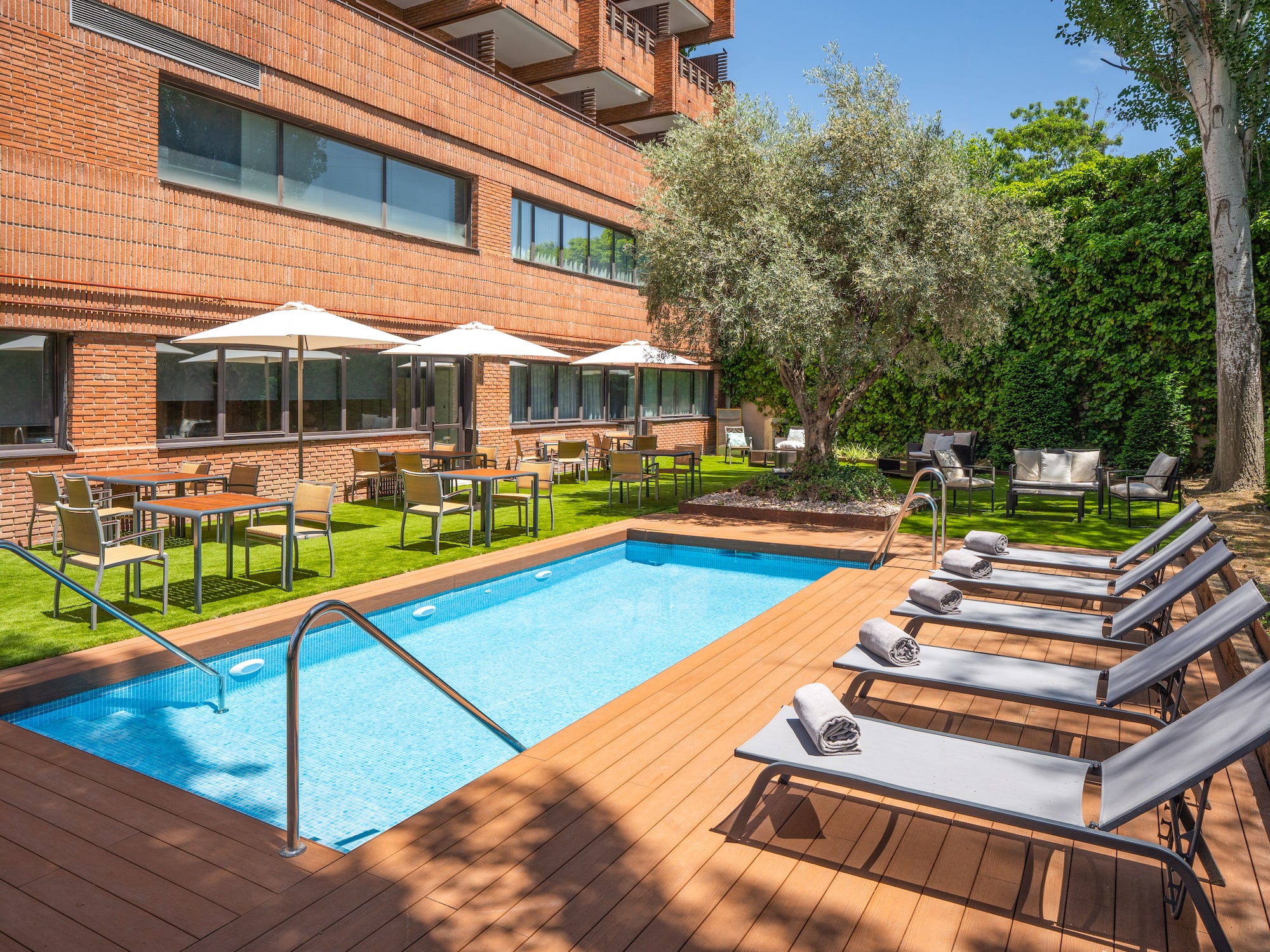 Outdoor hotel swimming pool with wooden deck, sun loungers, and garden dining.
