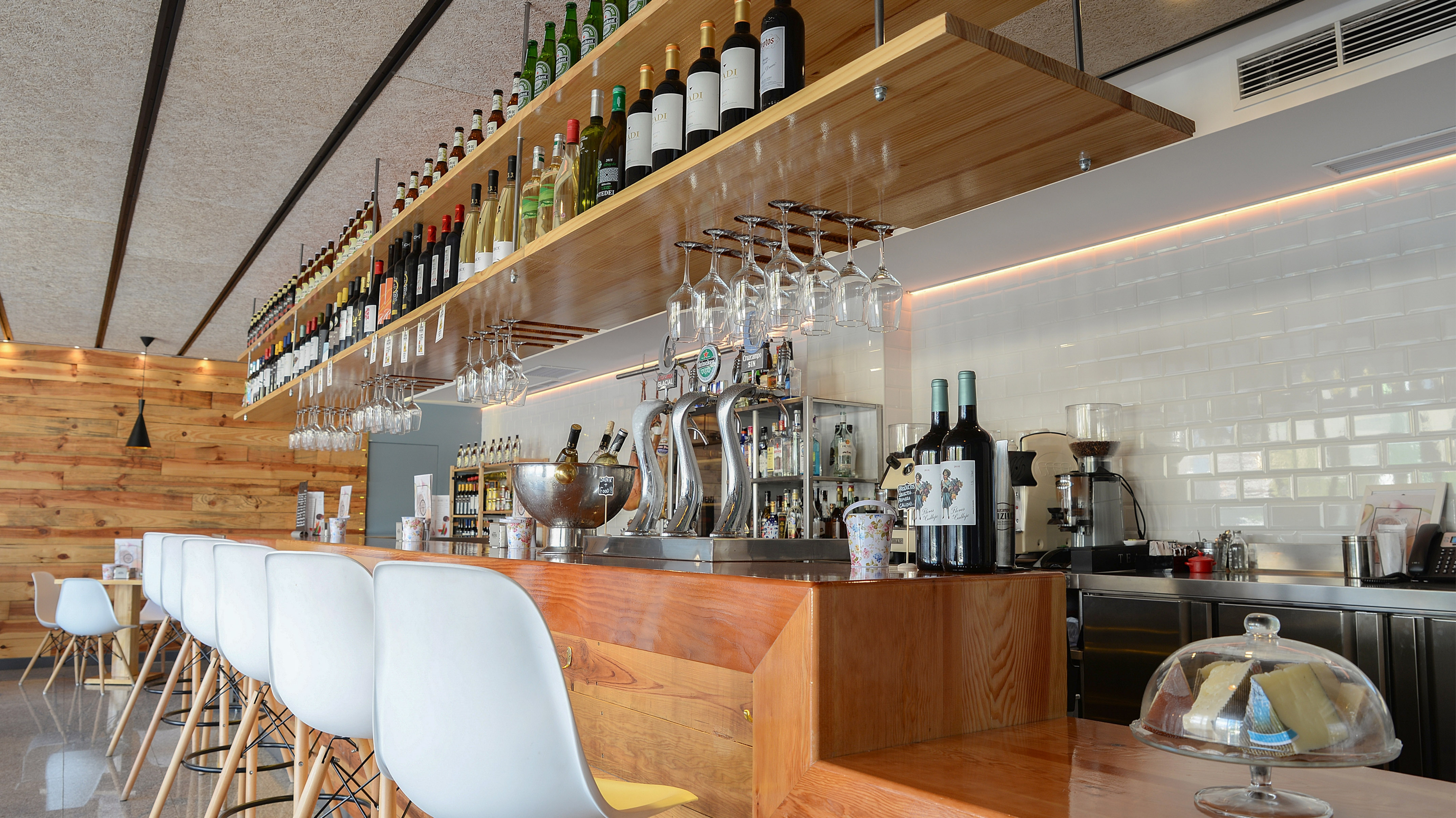 Stylish bar with wooden counter, white stools, wine bottles, and beer taps.