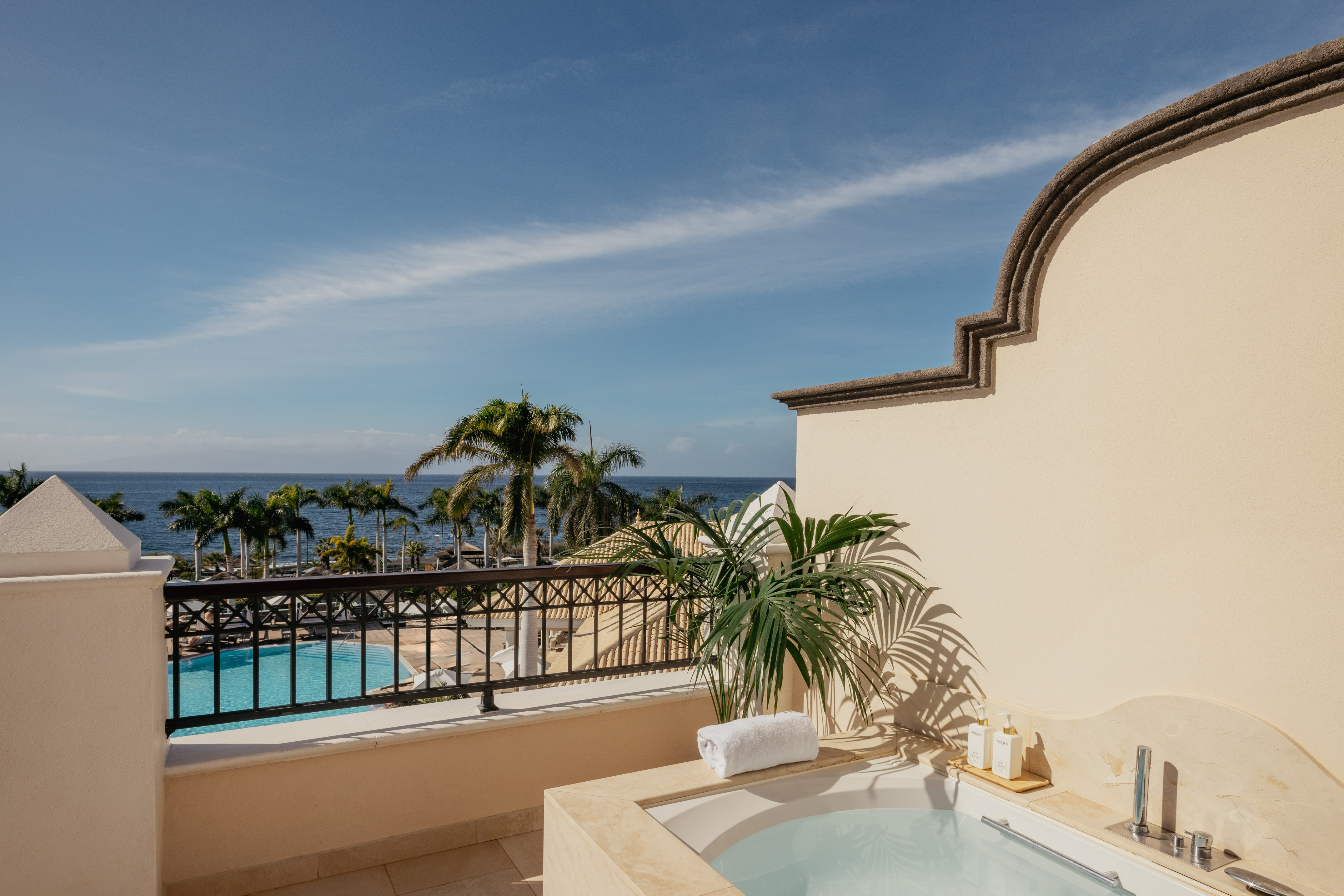 Hotel balcony with outdoor hot tub overlooking ocean, pool, and palm trees.