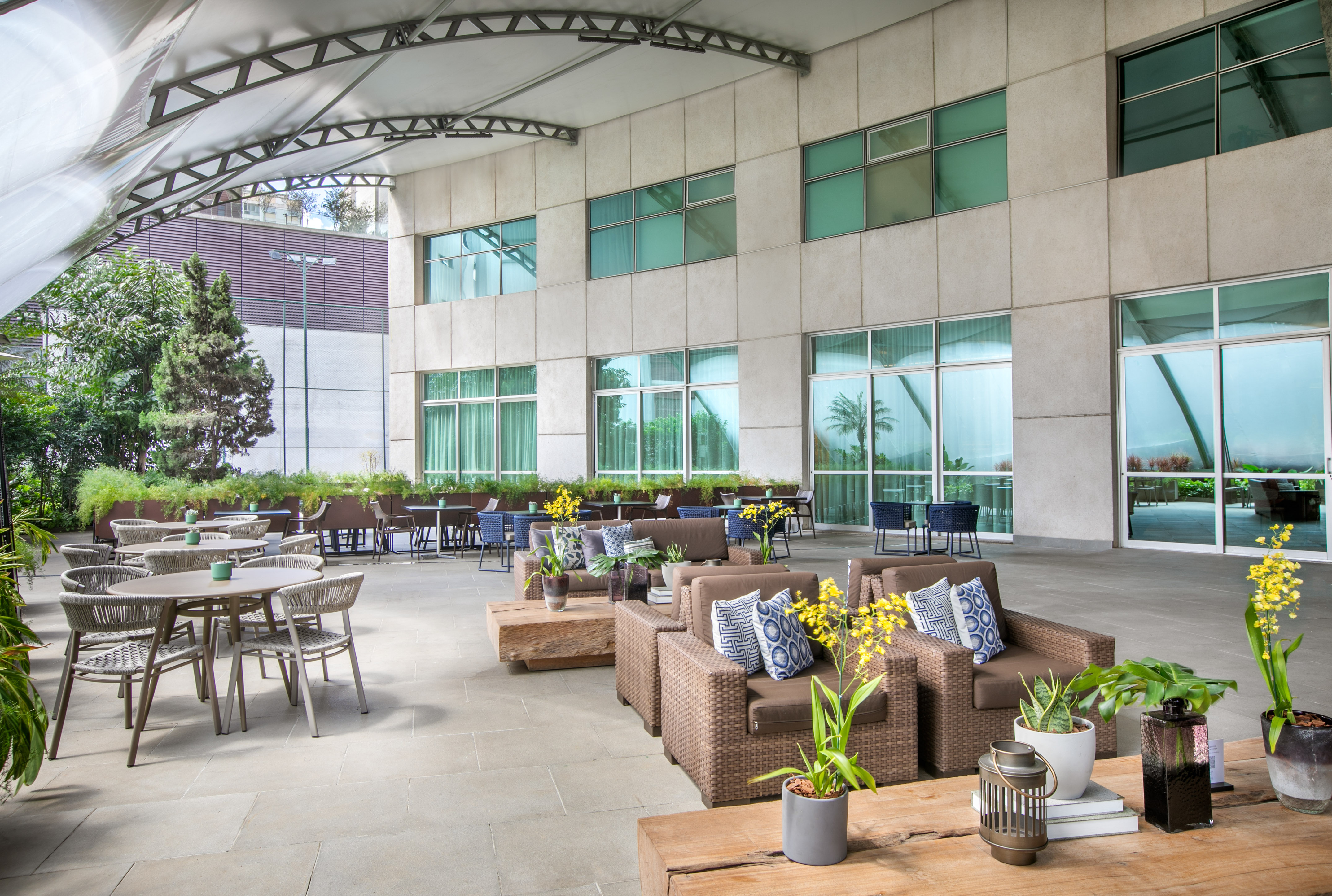 Modern hotel patio with rattan lounge and dining sets, green plants.