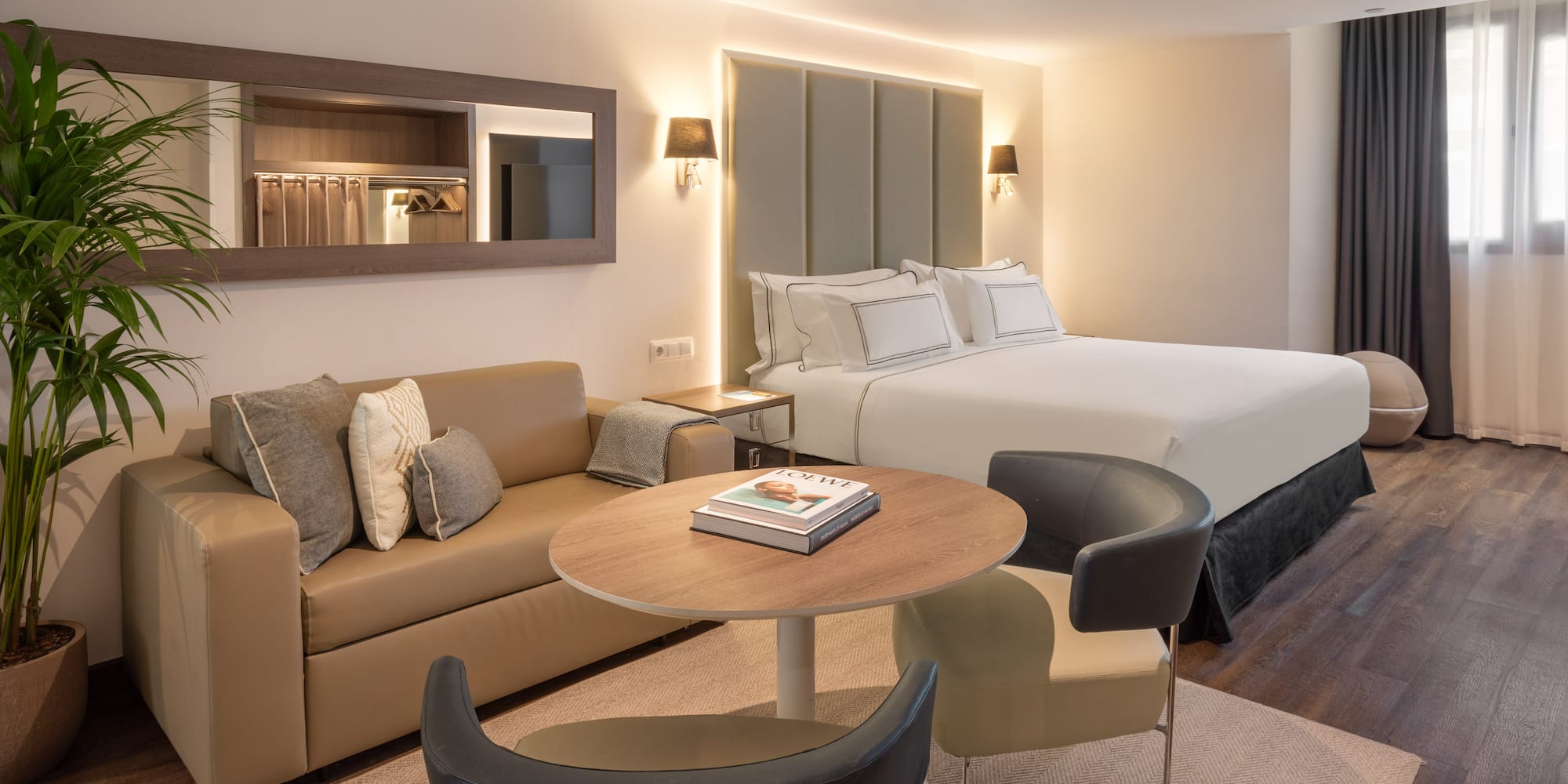 Modern hotel suite with bed, lounge area, coffee table, and warm lighting.