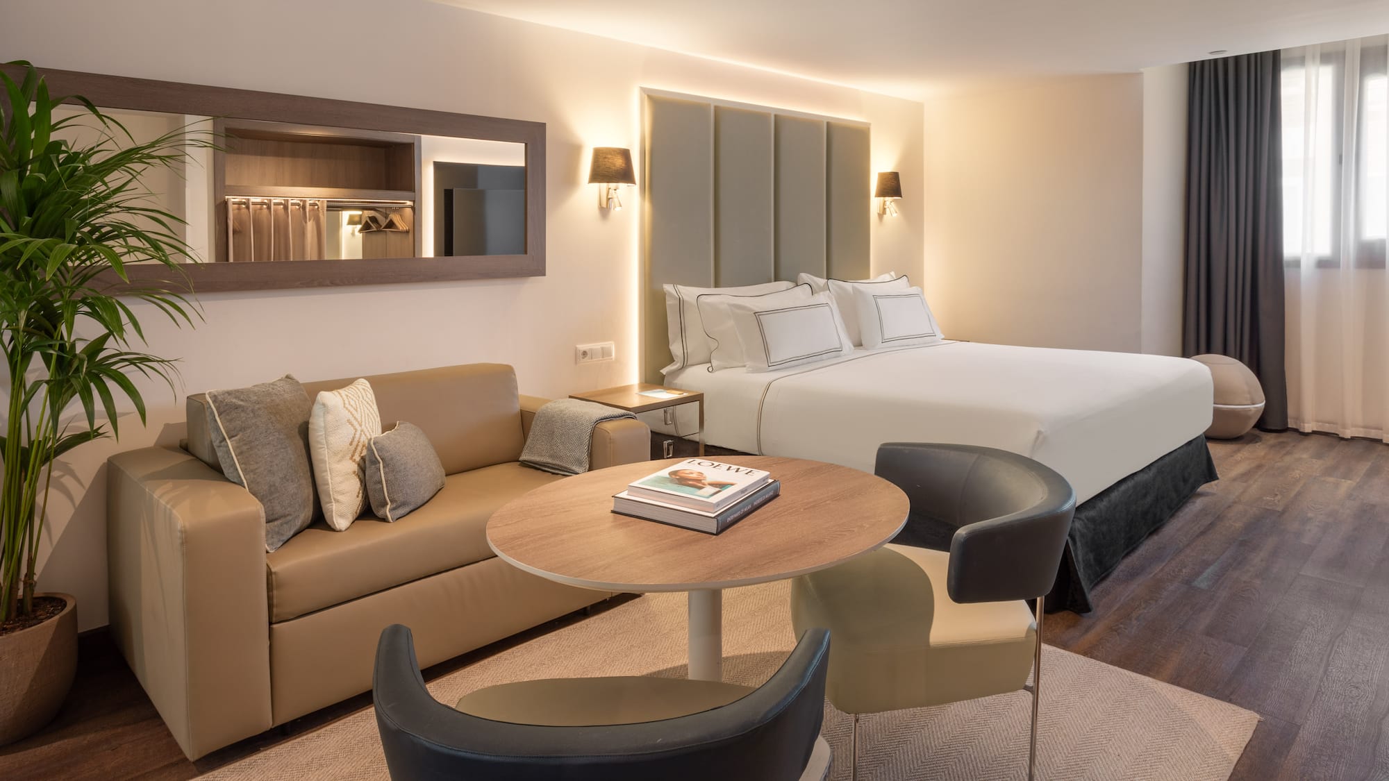 Modern hotel suite with bed, beige sofa, coffee table, and wall mirror.