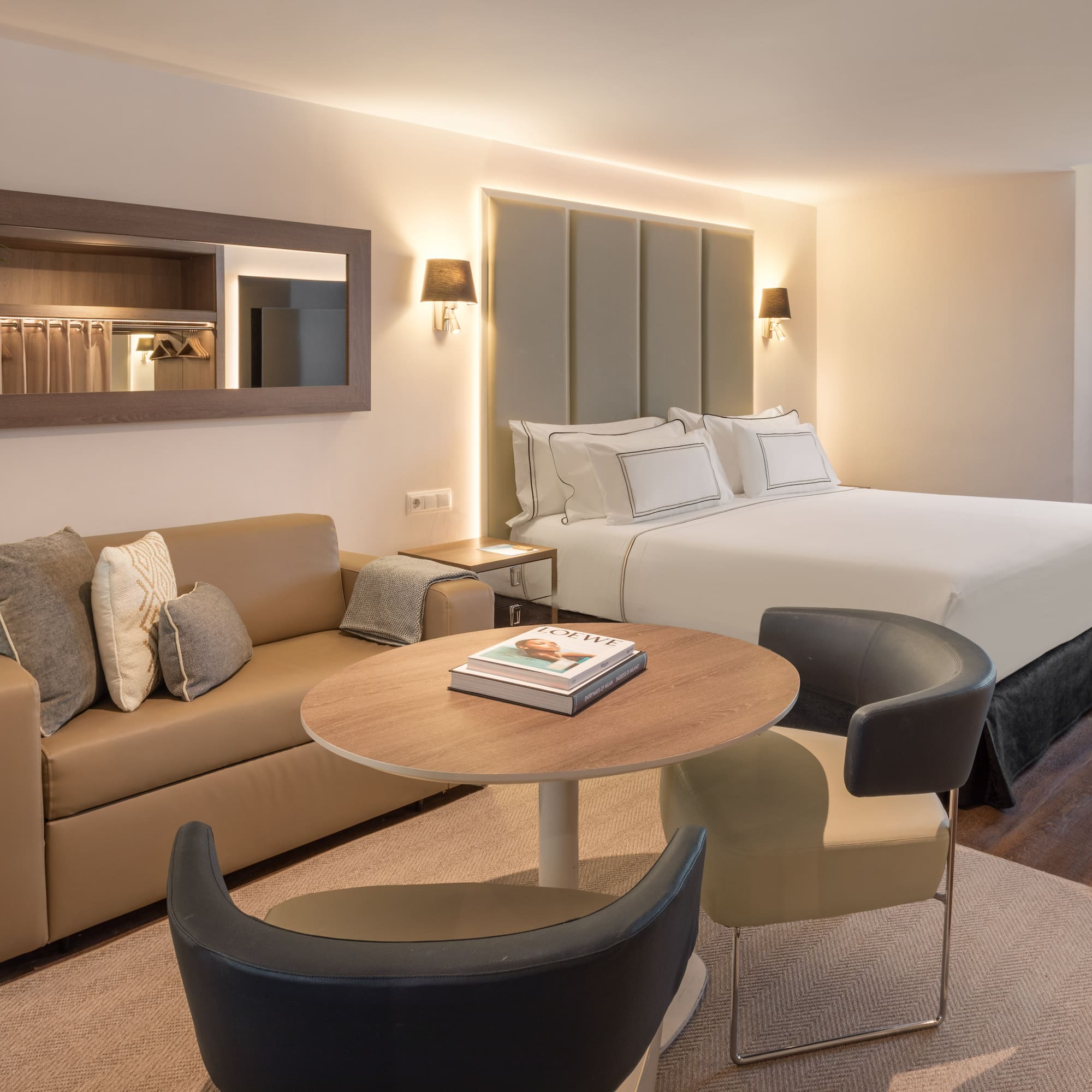 Modern hotel suite with bed, beige sofa, coffee table, and wall mirror.