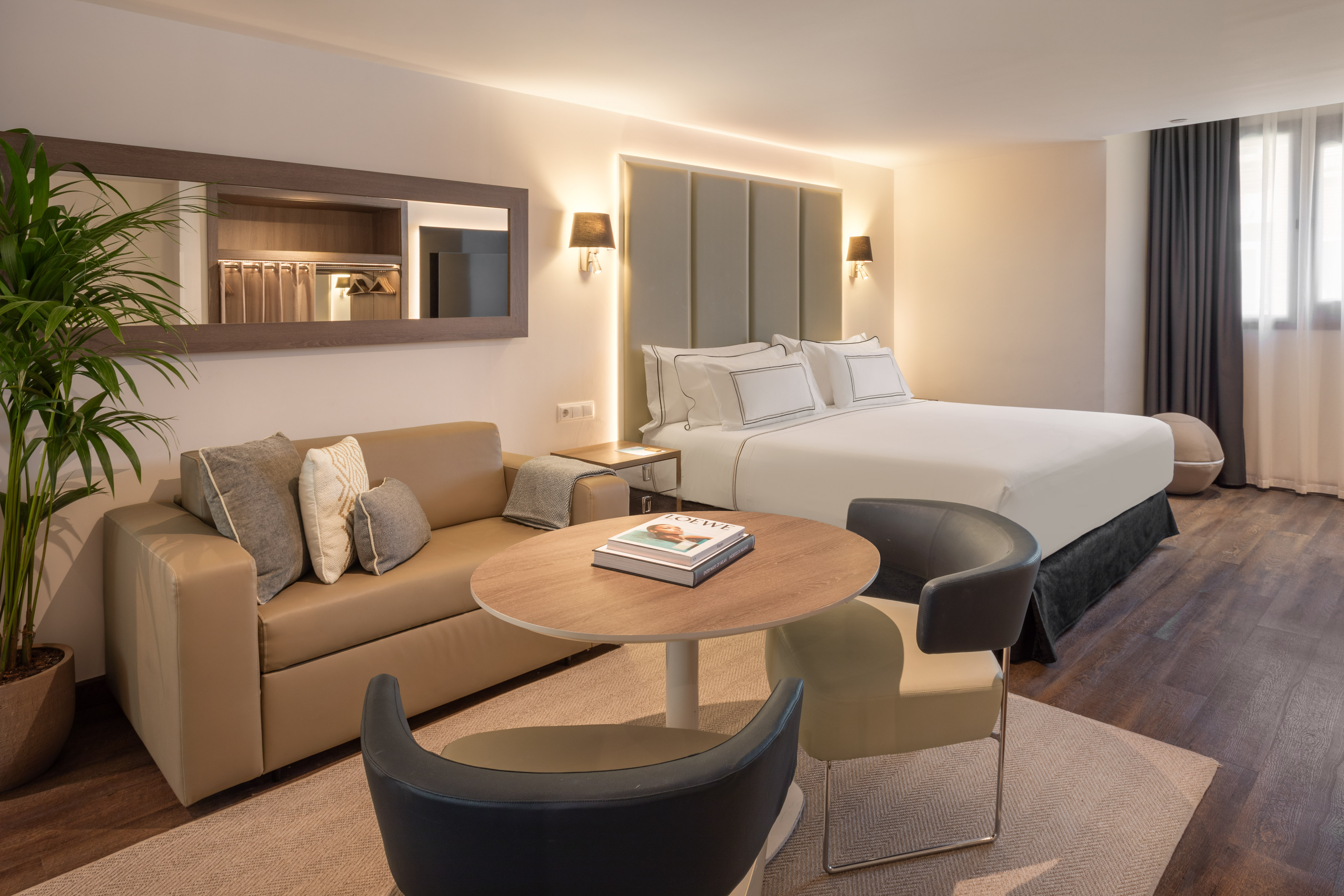 Modern hotel suite with bed, beige sofa, coffee table, and wall mirror.
