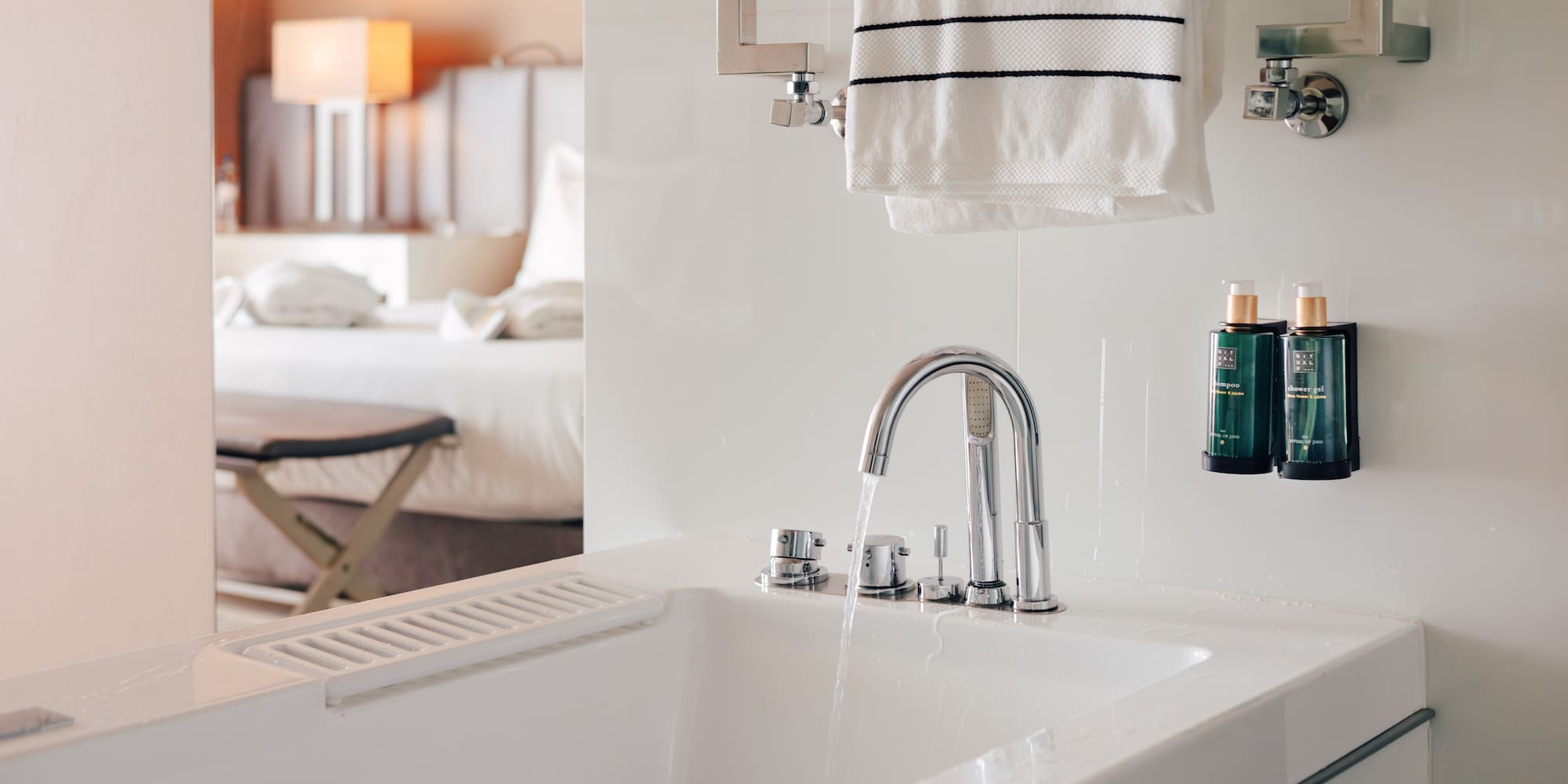 Modern hotel bathroom with bathtub, running water, towels, and amenities.