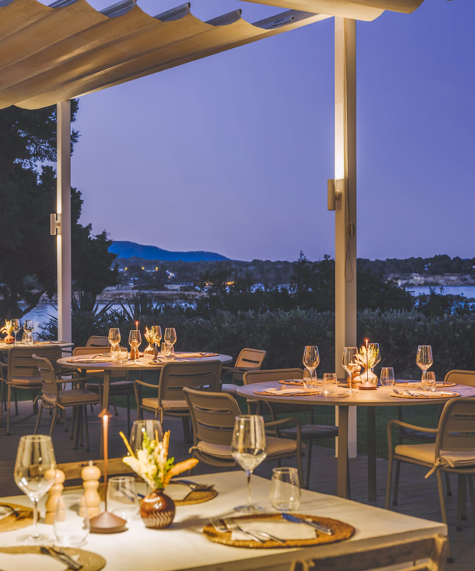 Waterfront outdoor dining patio, tables set for dinner at dusk, sea view.