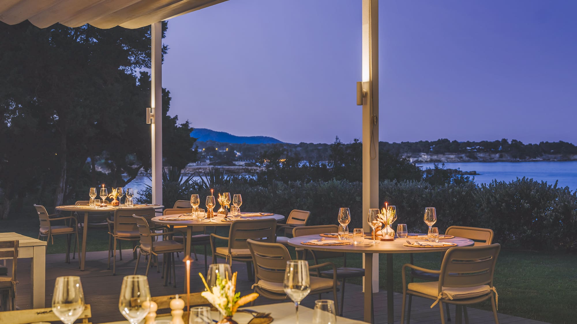 Outdoor restaurant patio at dusk with set tables and a beautiful sea view.