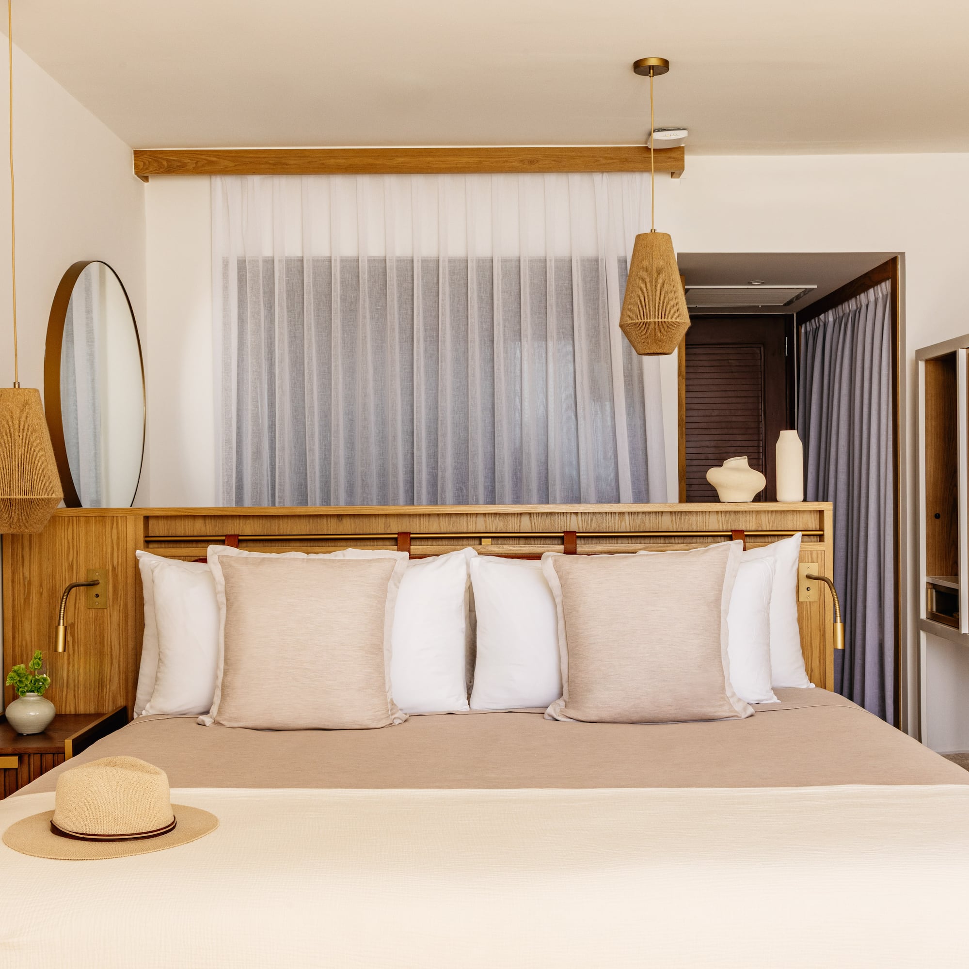 Modern hotel bedroom with light wood furniture, neutral bedding, woven lights, straw hat.