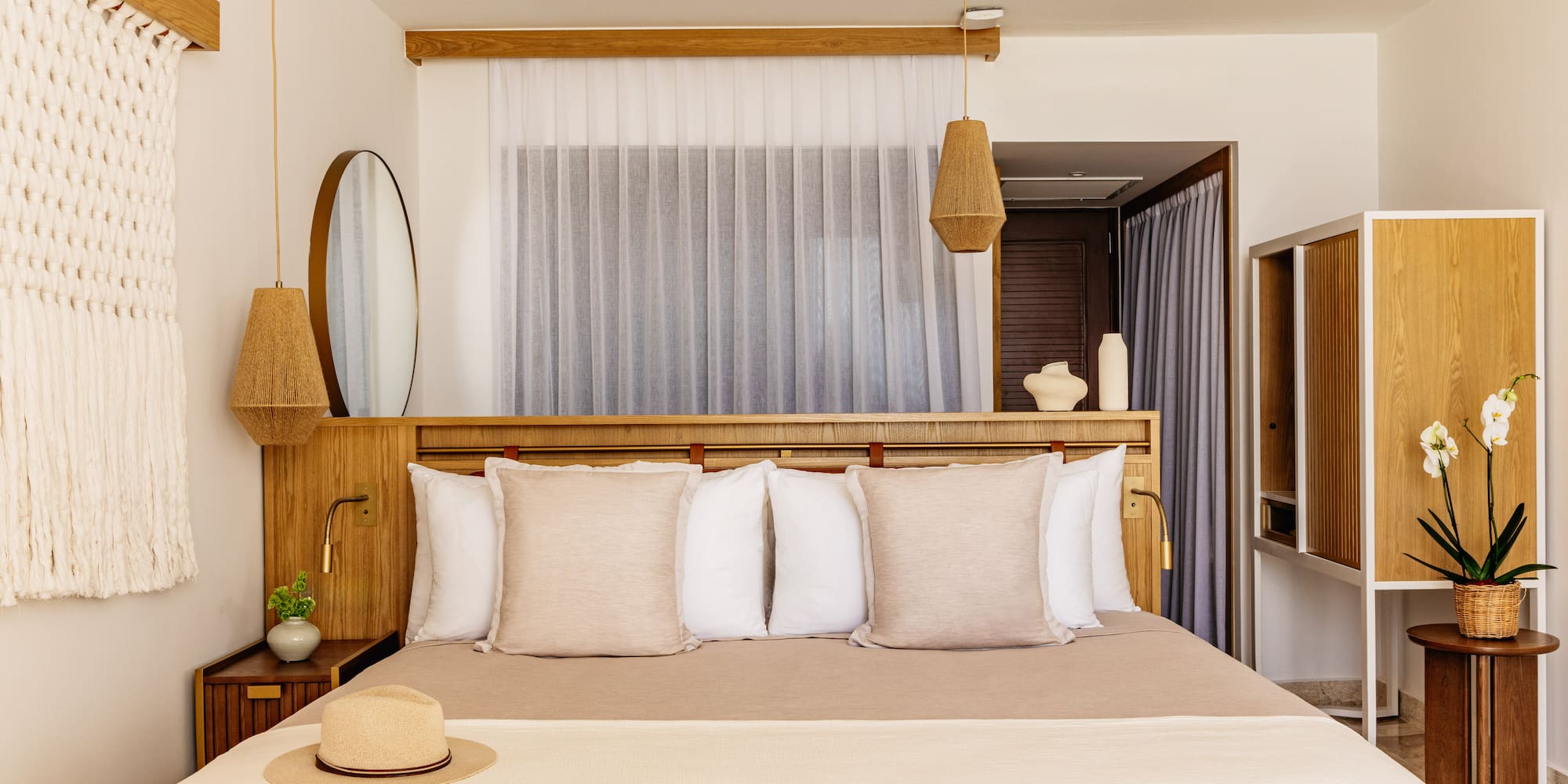 Modern hotel bedroom with light wood furniture, neutral bedding, woven lights, straw hat.