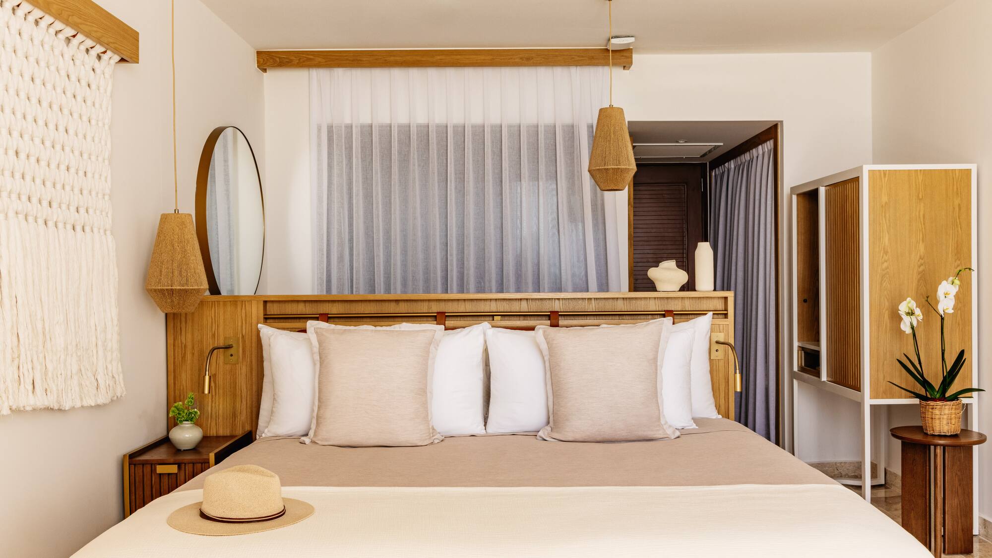 Modern hotel bedroom with light wood furniture, neutral bedding, woven lights, straw hat.