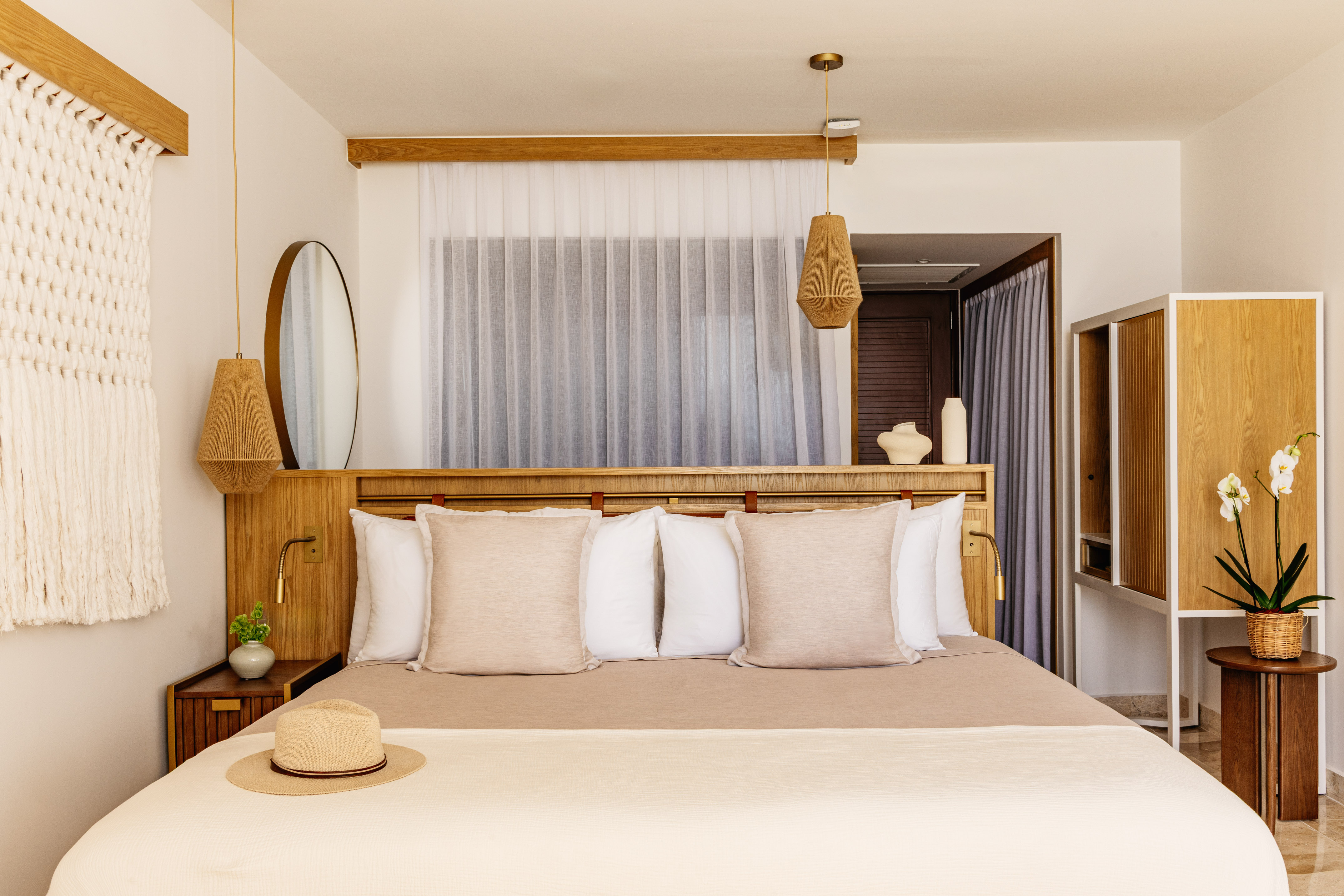 Modern hotel bedroom with light wood furniture, neutral bedding, woven lights, straw hat.