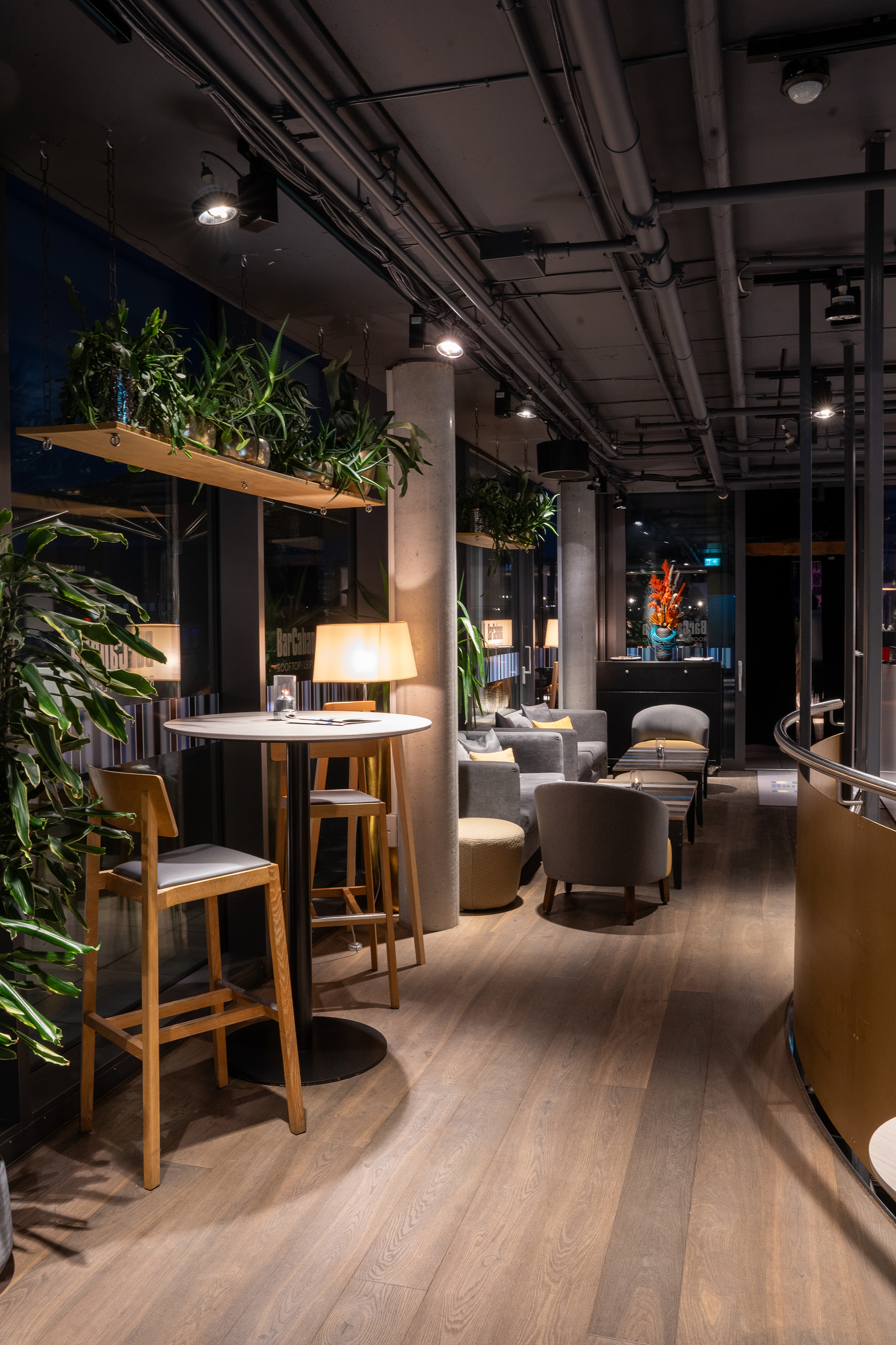 Cozy modern bar lounge with industrial ceiling, plants, warm lighting, and wood floor.