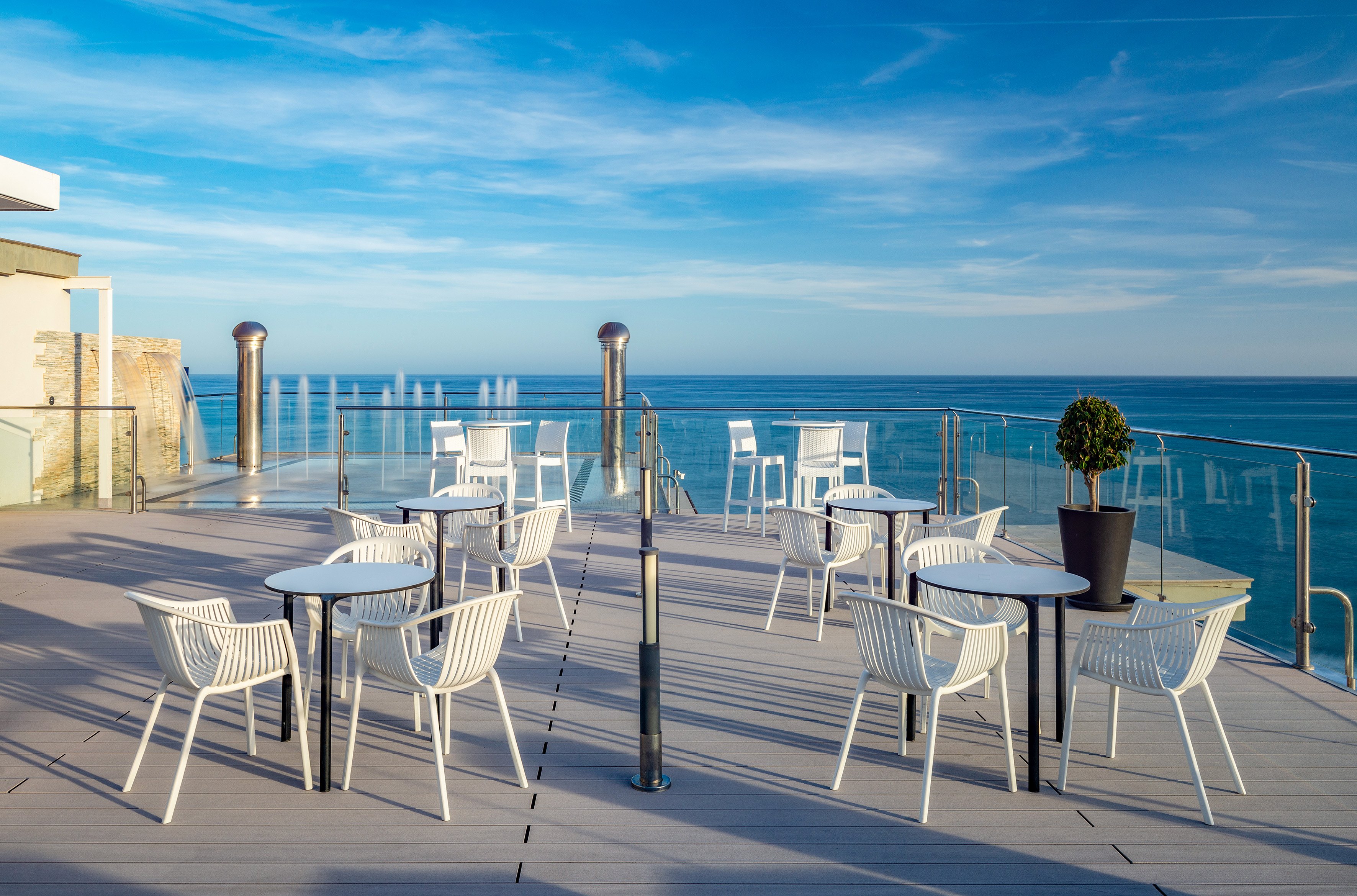 Sunny outdoor cafe terrace with sea view, white furniture, and fountains.