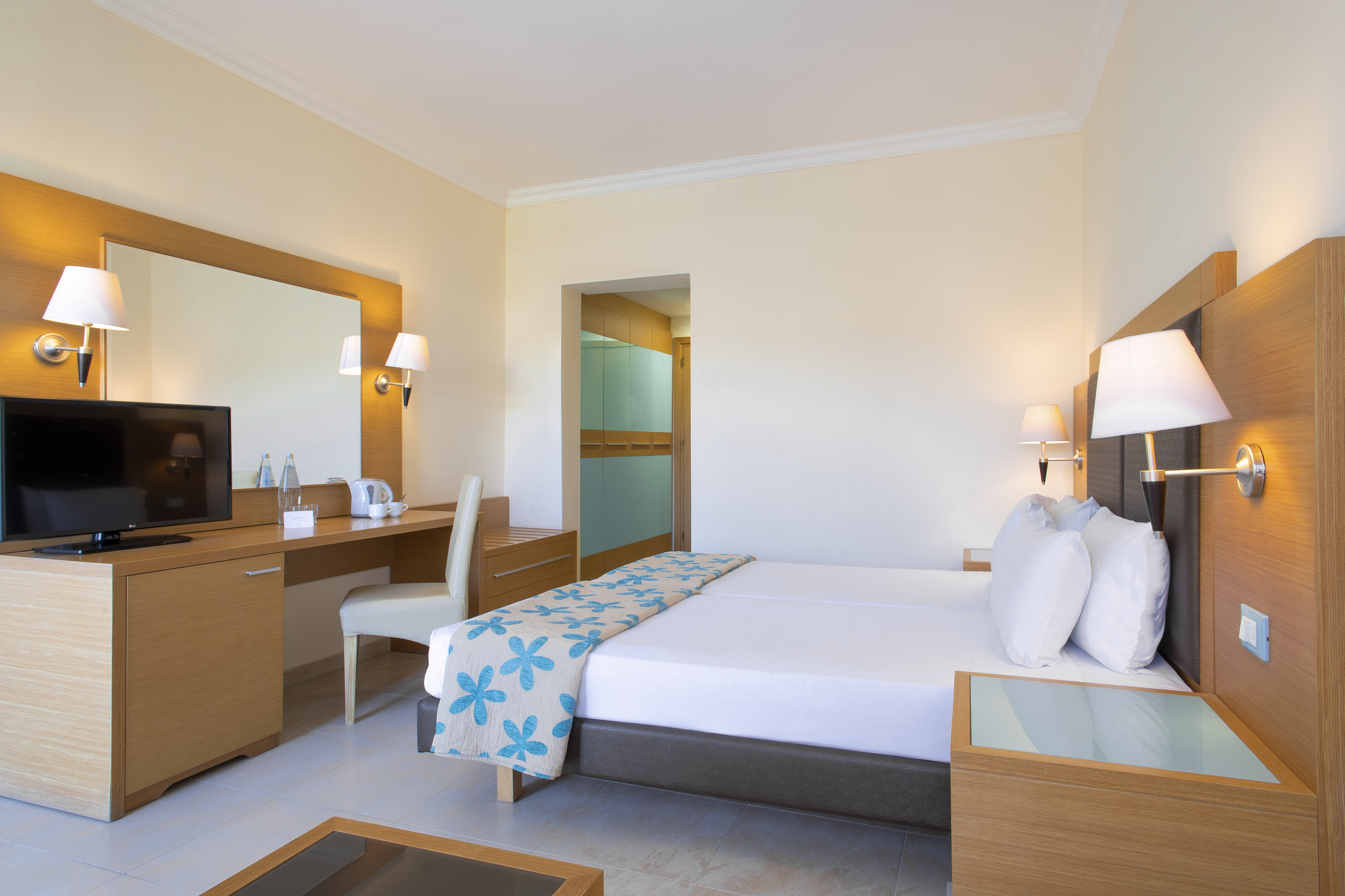 Hotel bedroom with twin beds, blue floral bed runner, wooden desk, TV, and mirror.