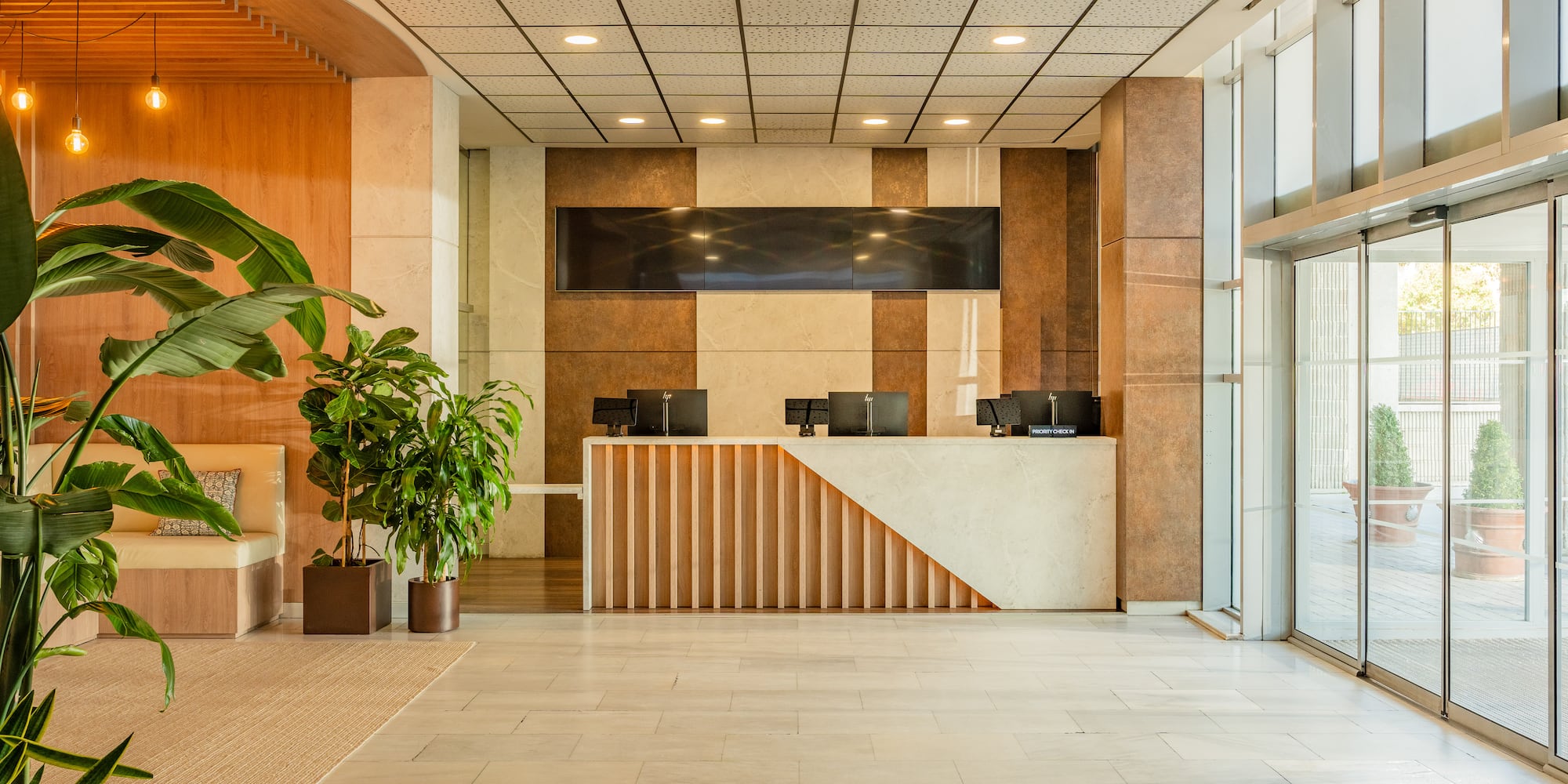 Modern hotel lobby with reception desk, lush plants, and glass doors.