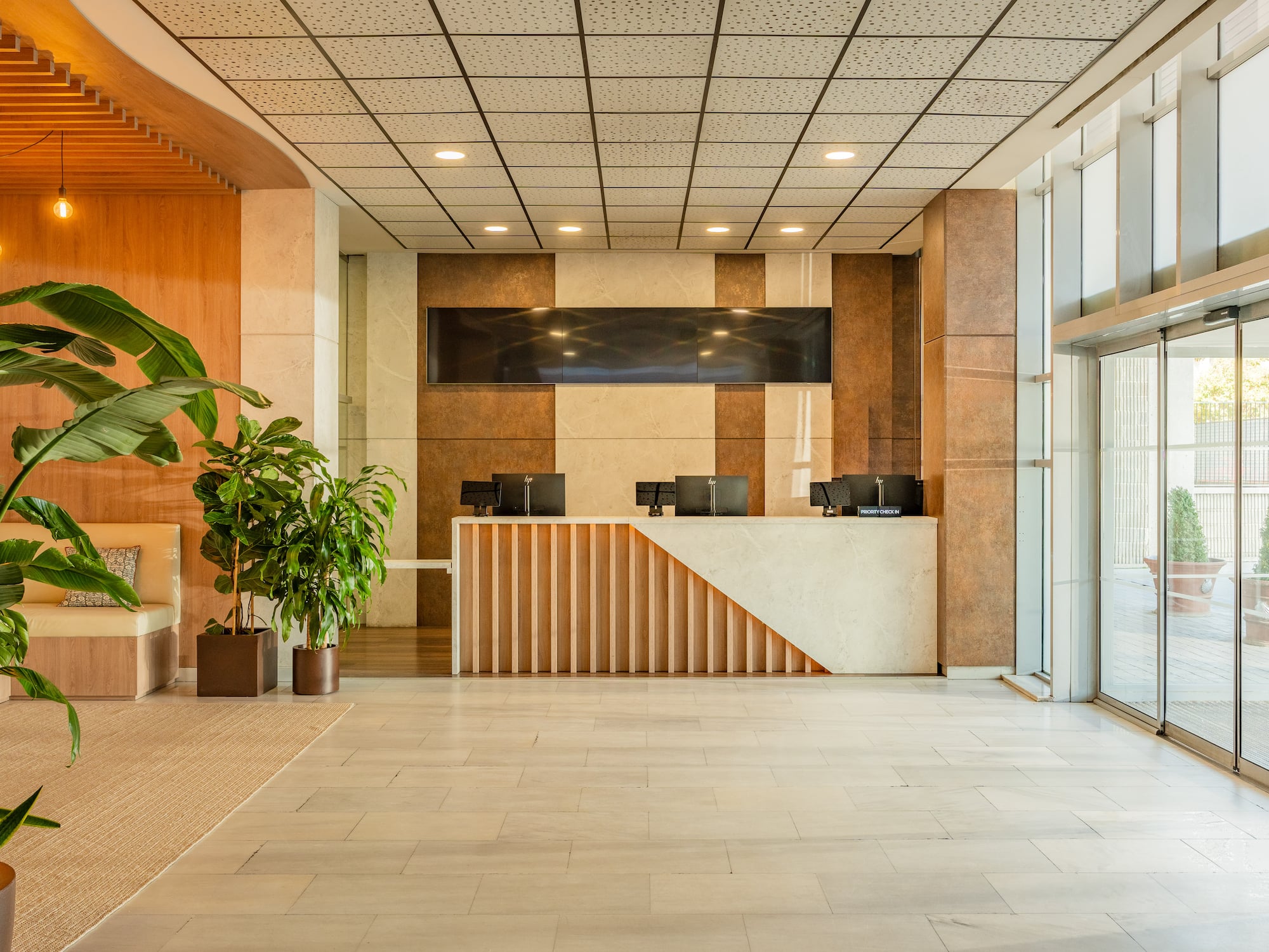 Modern hotel lobby with reception desk, lush plants, and glass doors.